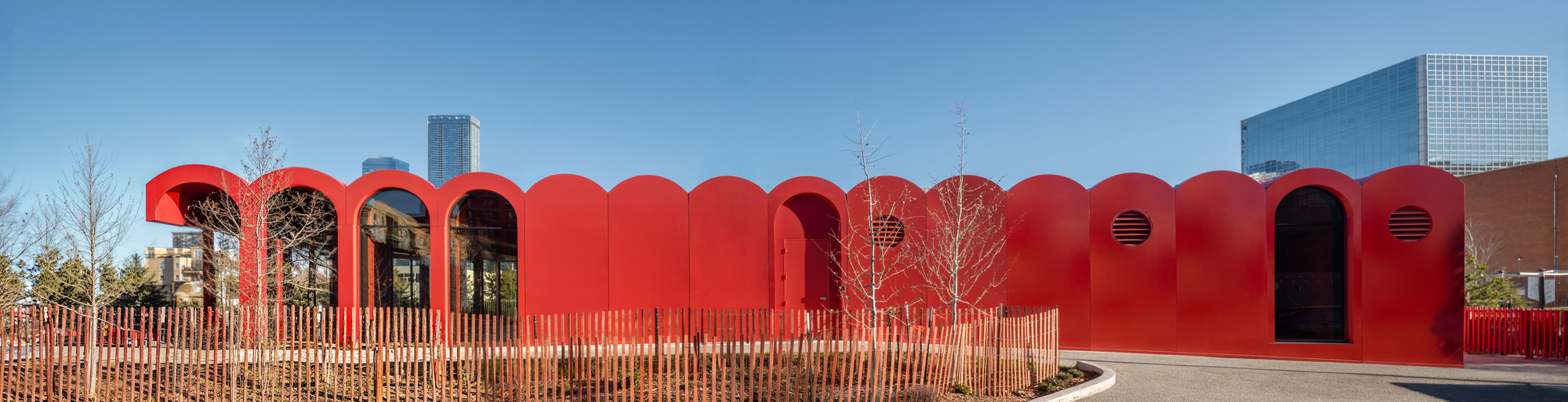 Long red facade with repeating arched profiles and timber fencing along the entry plaza