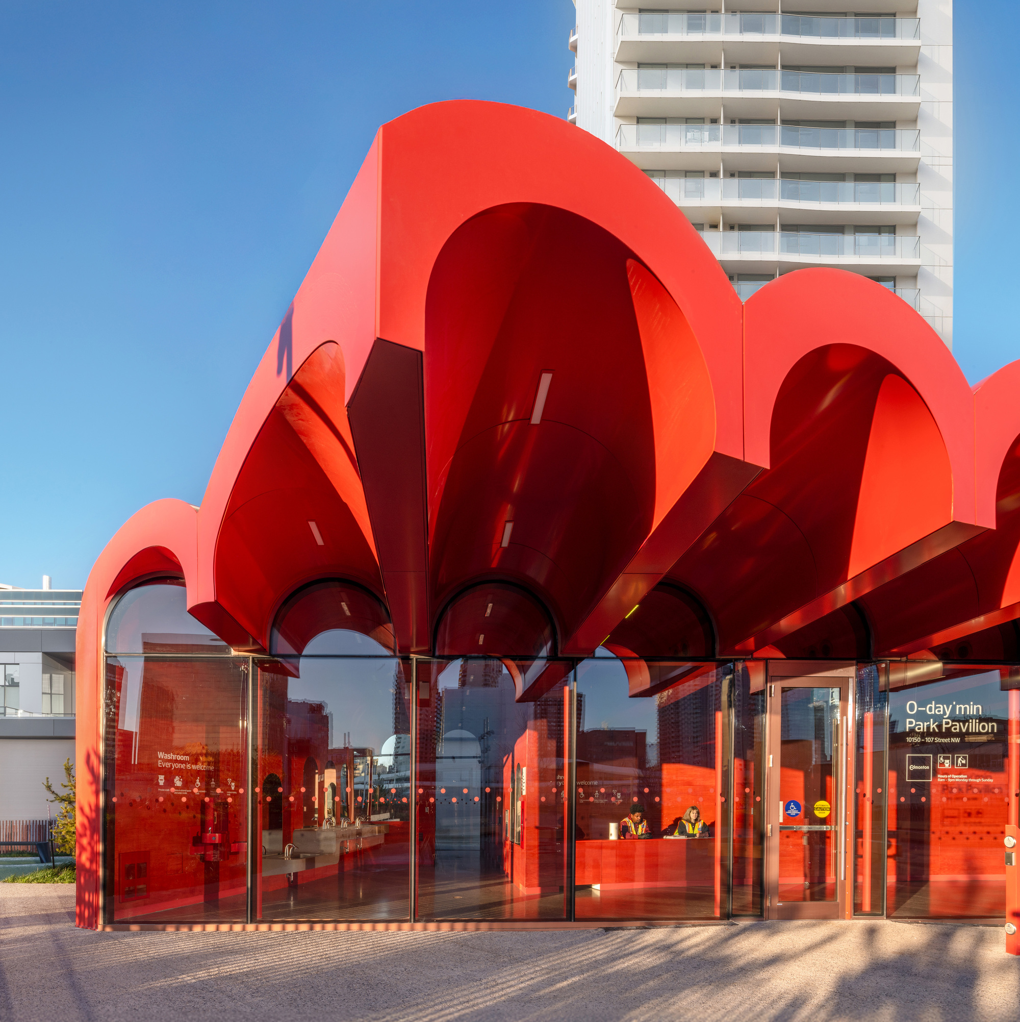 Glass-walled entrance pavilion beneath radiating red arched roof at twilight with people inside