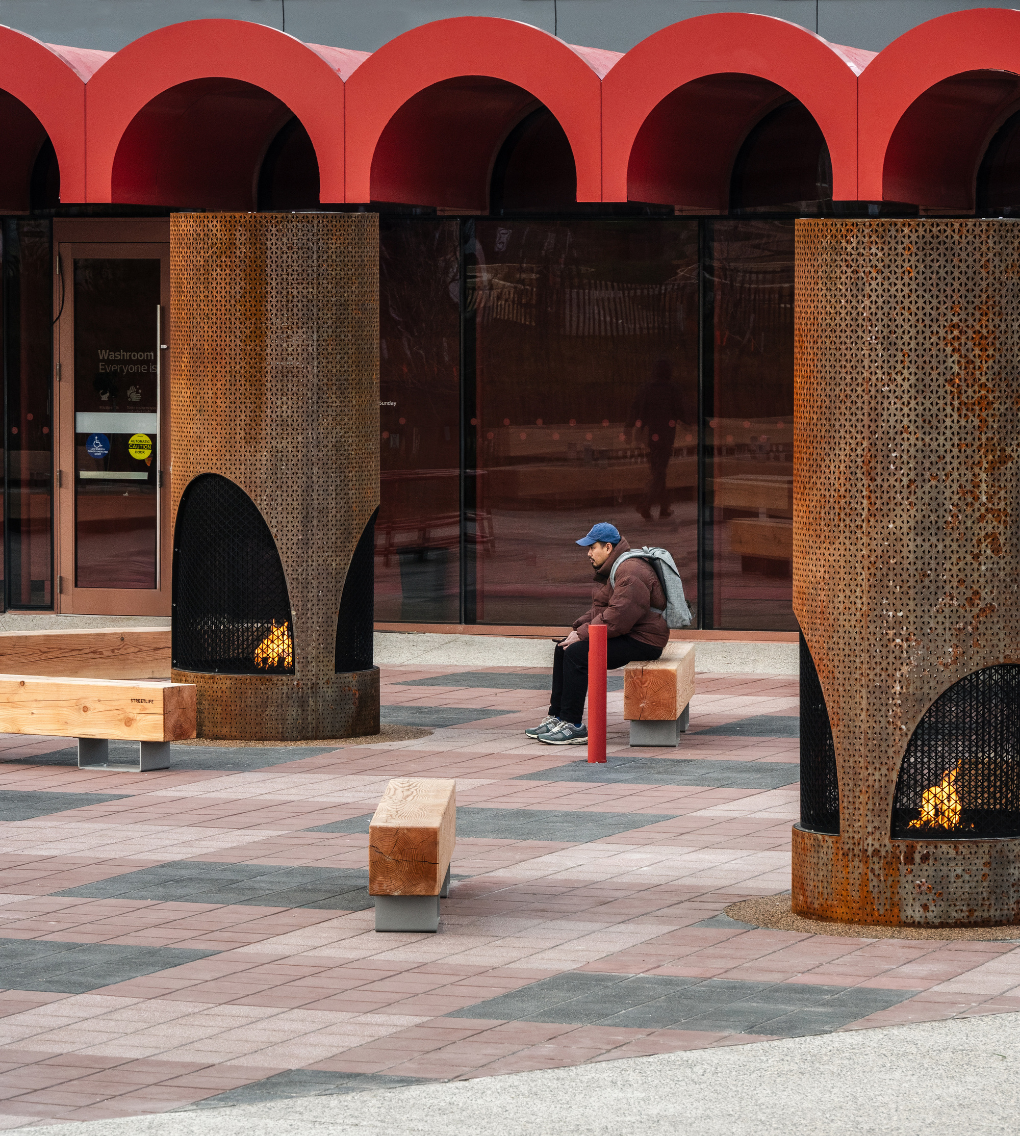 Perforated metal heating towers on the tiled plaza with a visitor seated on a timber cube