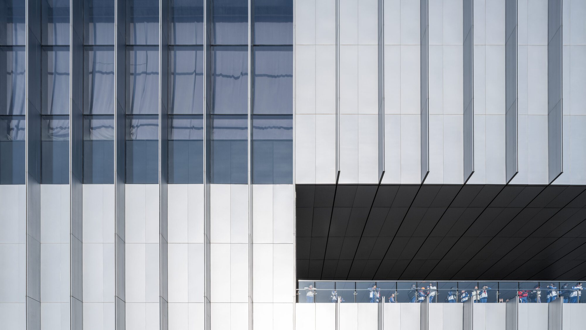 Facade detail showing white vertical panels meeting black soffit with glazed gallery strip revealing visitors inside