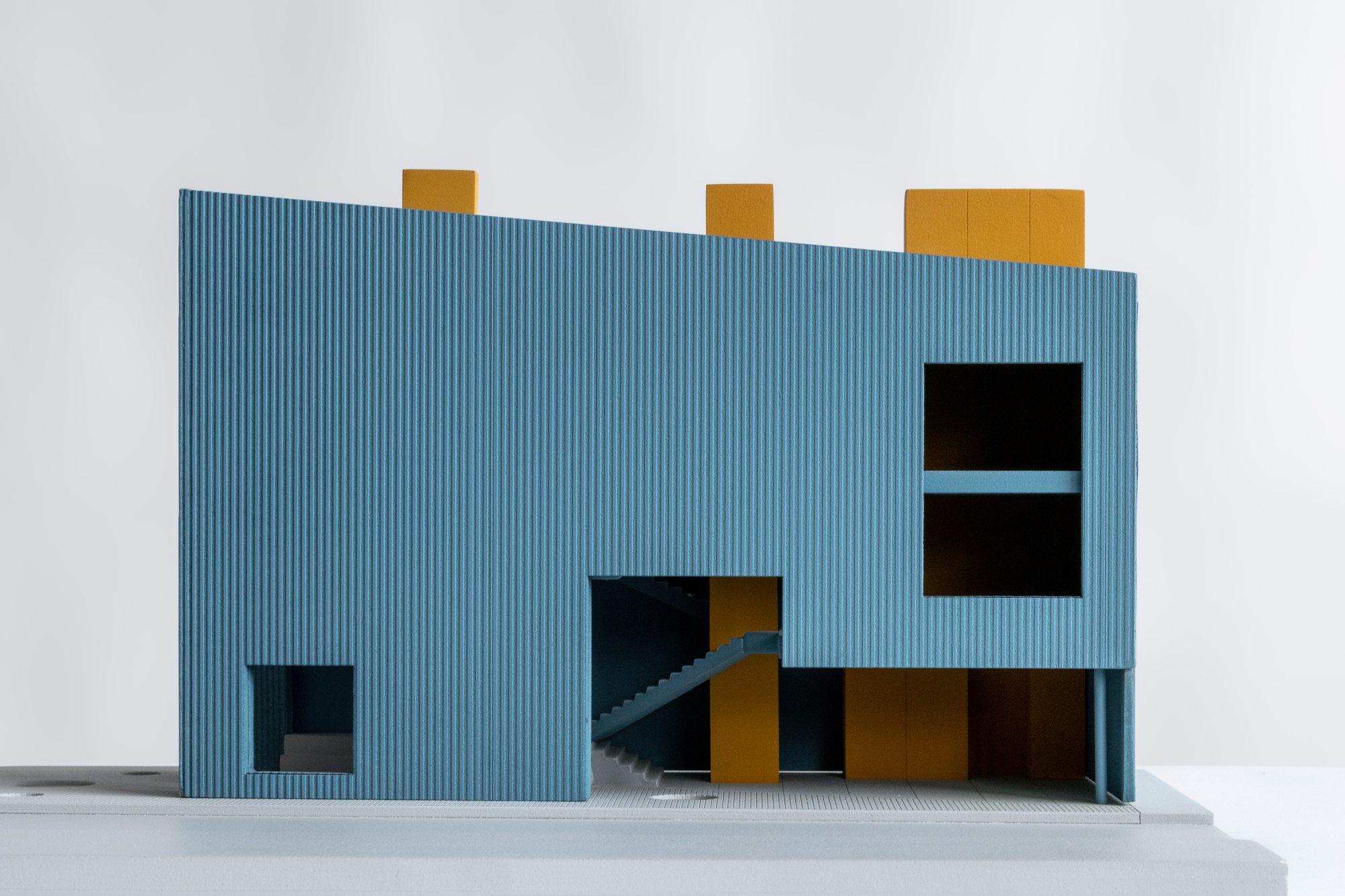 Physical model with corrugated blue cladding and yellow volumetric elements puncturing the facade