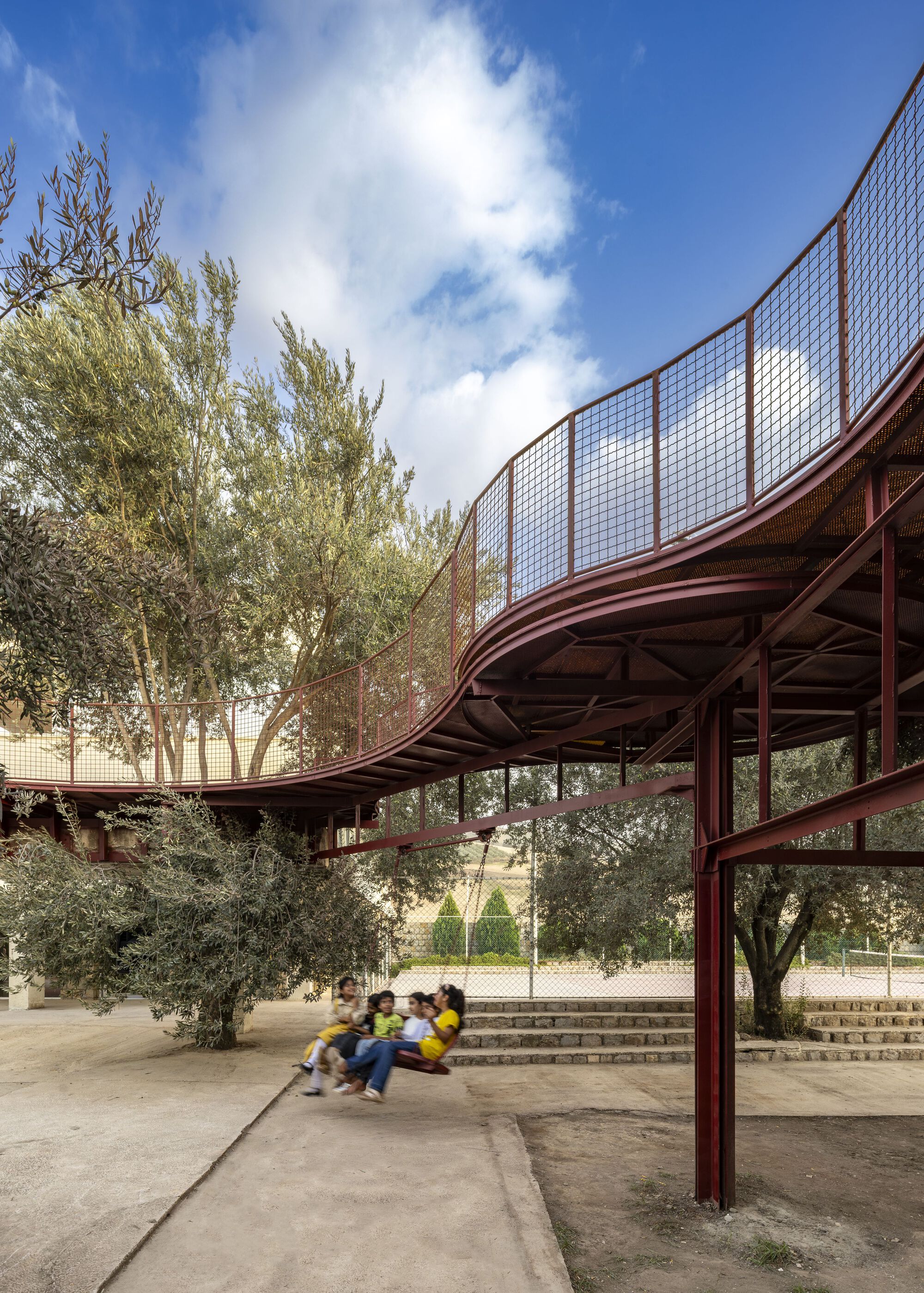 Curving canopy with mesh screens and red steel columns sheltering a stepped amphitheater among olive trees