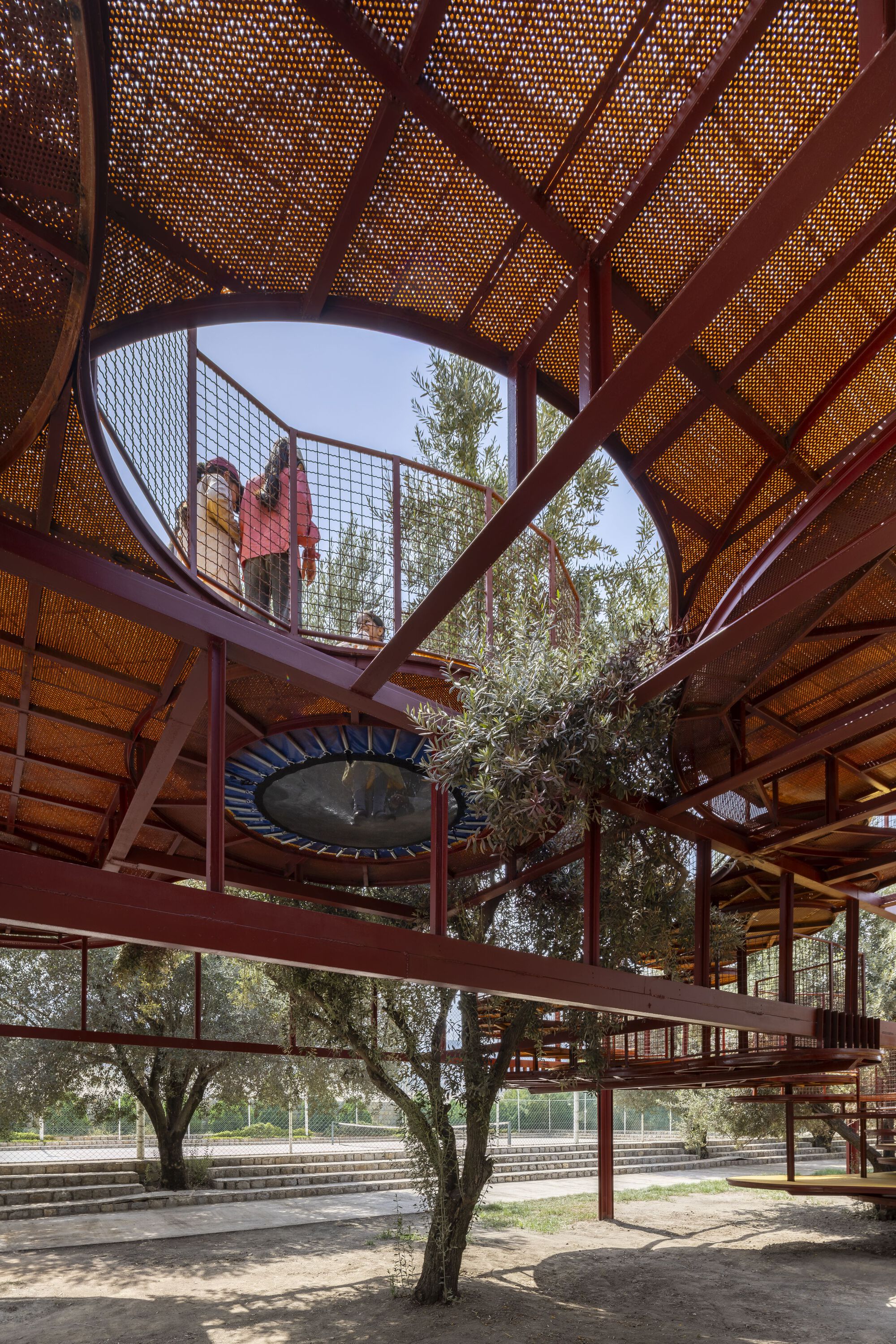 Upward view through perforated metal ceiling panels to an upper walkway with mesh railings and trees