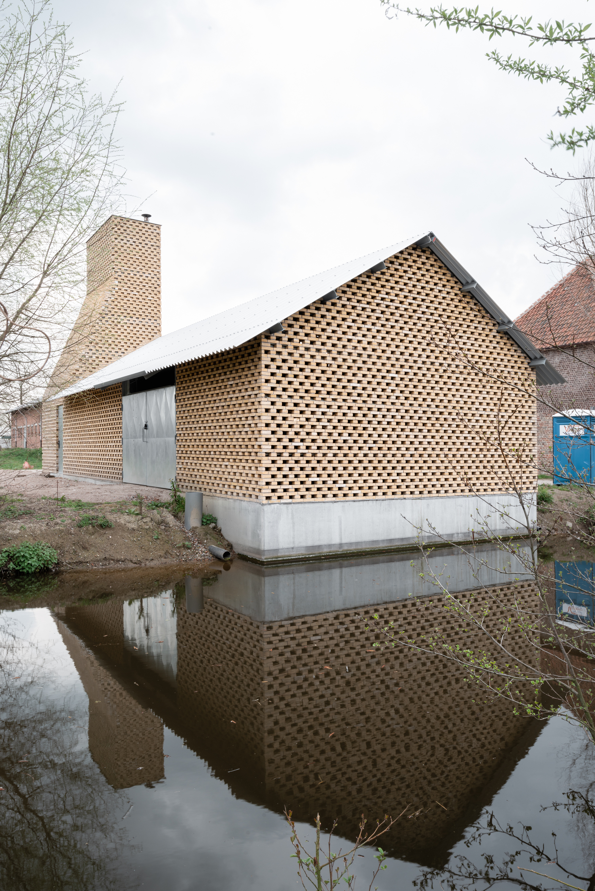 Gabled volume with perforated brick facade on concrete base reflected in adjacent canal water
