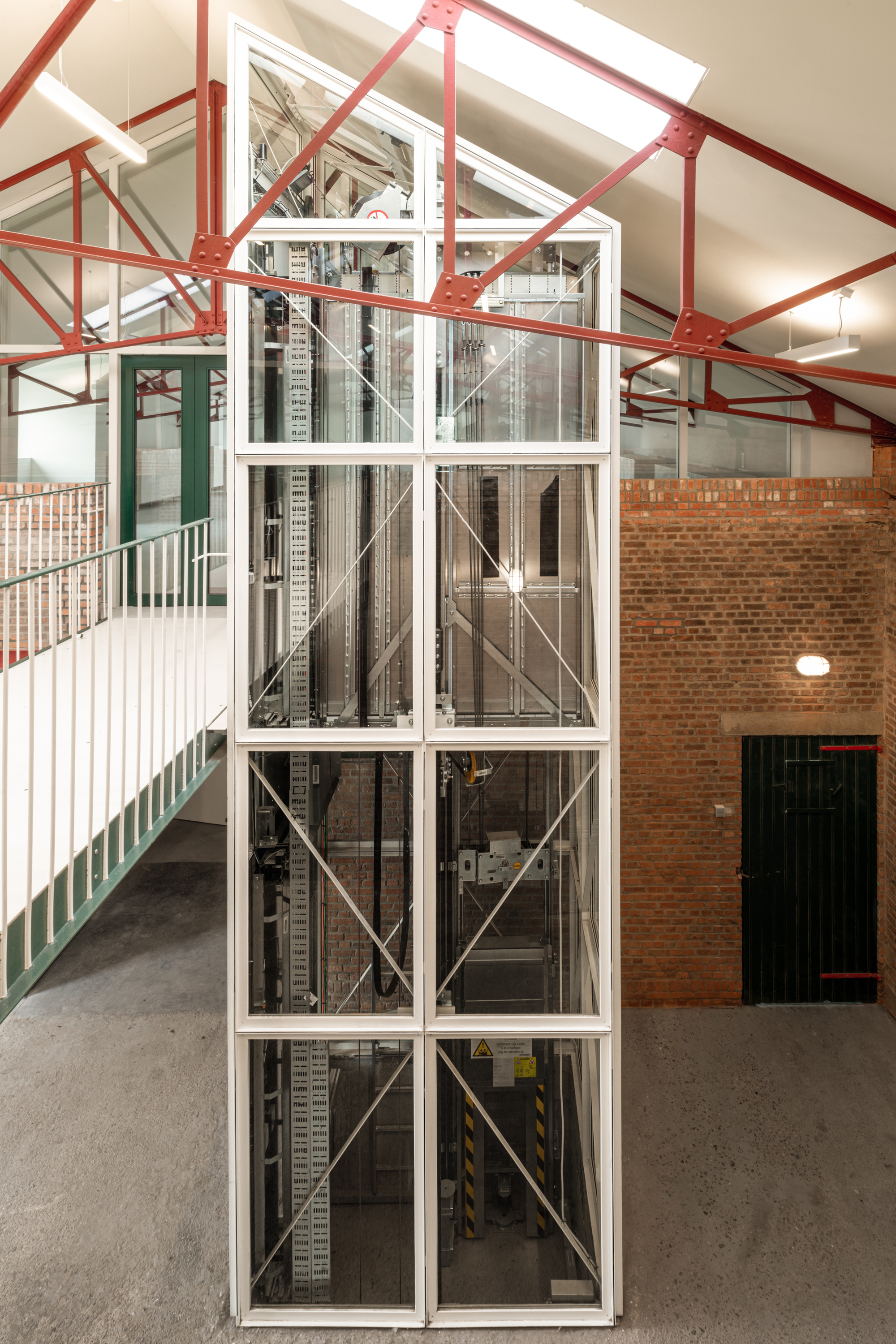 Glass-enclosed elevator shaft with exposed steel cross-bracing inside a warehouse with red painted trusses
