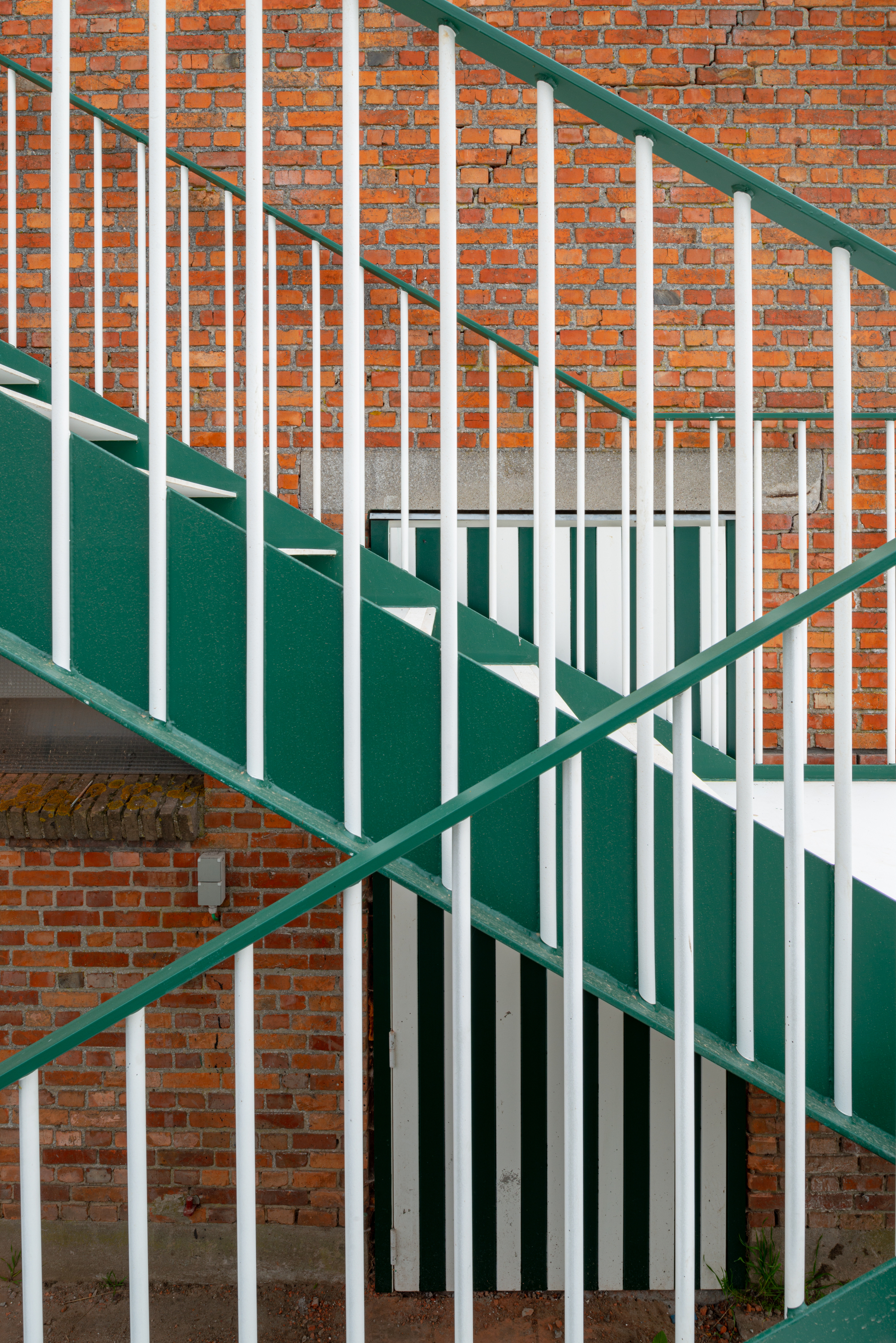 Green-and-white painted steel staircase with vertical bar railings ascending against exposed brick