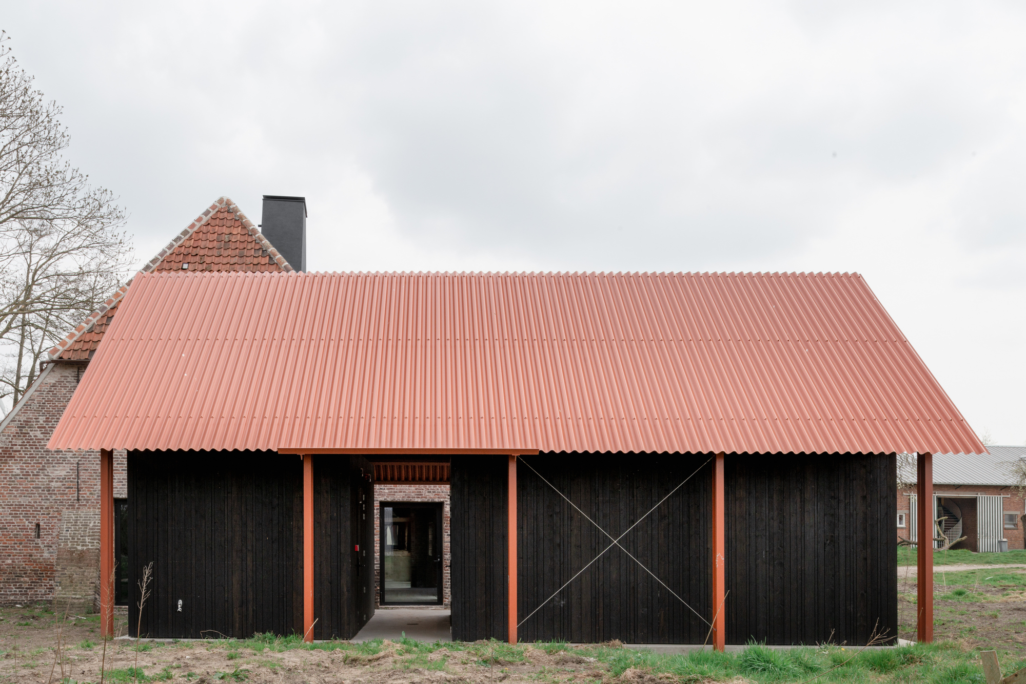 Black timber-clad barn volume with coral corrugated metal roof and orange steel columns under overcast sky