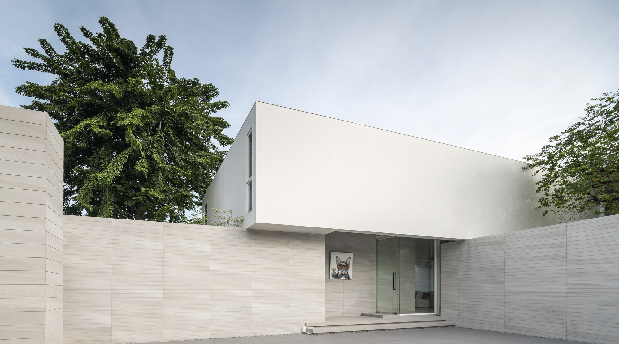Street view of the white upper volume floating above horizontal stone walls and mature trees