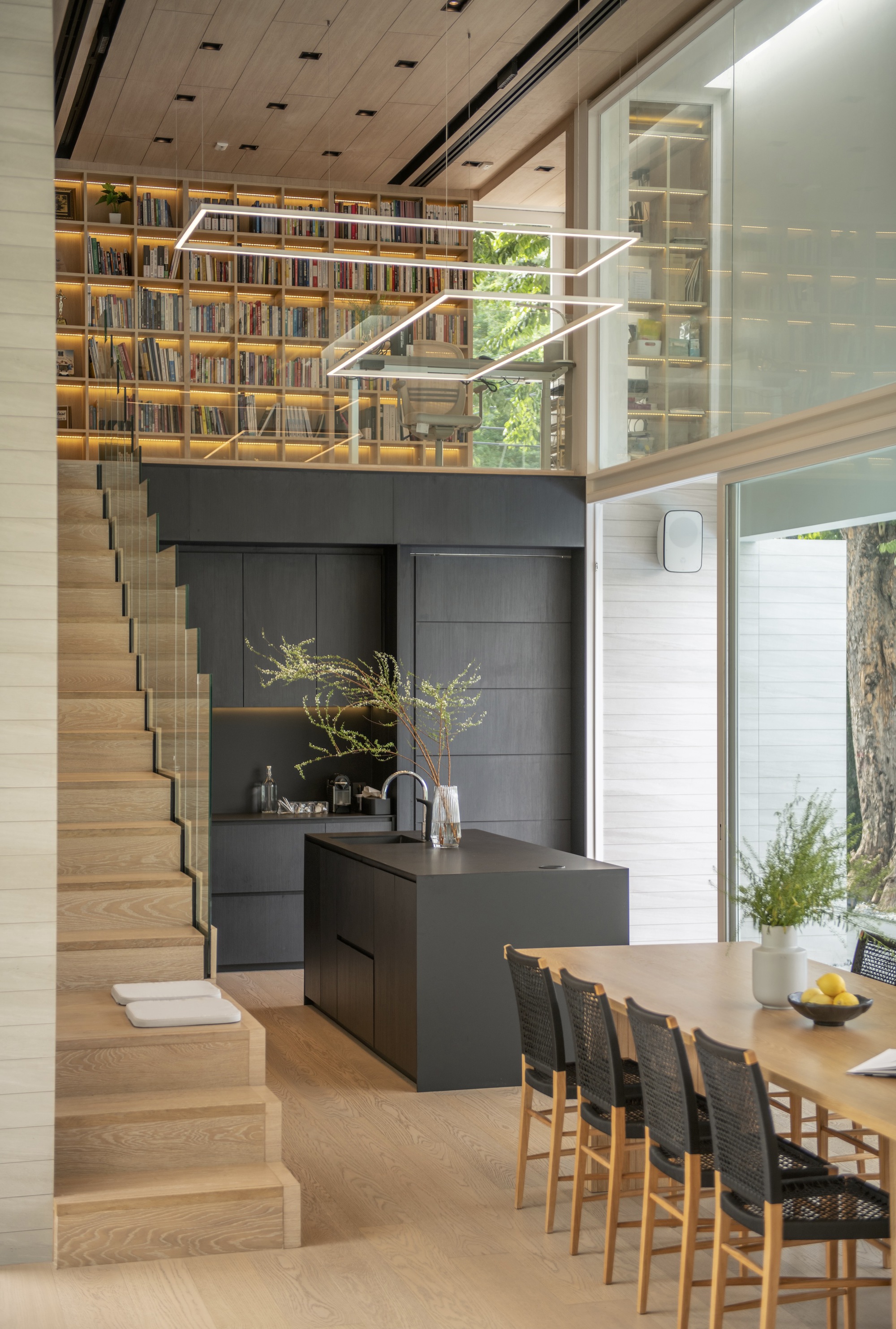 Double-height interior showing timber staircase ascending to mezzanine library above black kitchen island