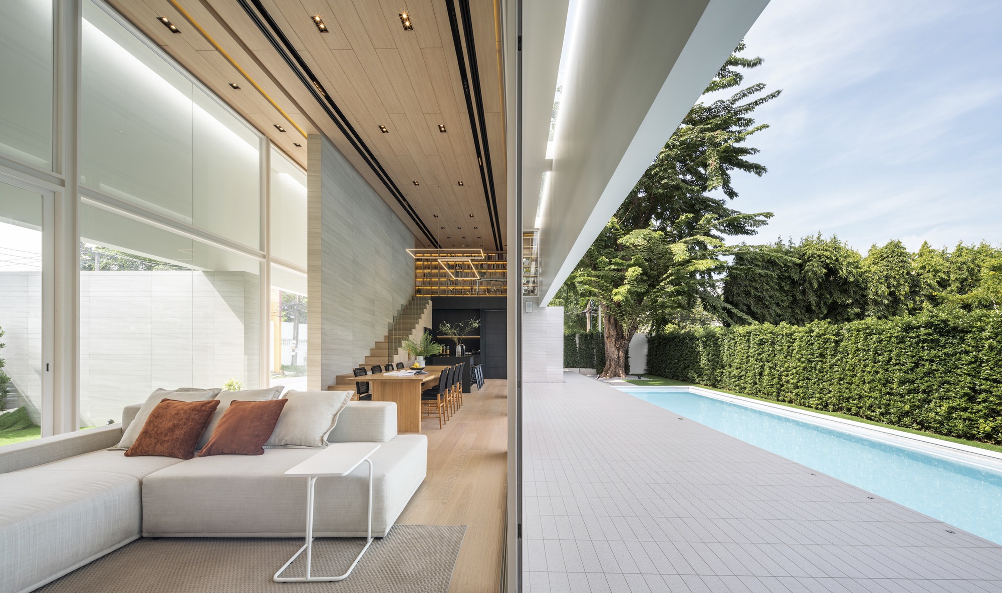 Open-plan living area with timber ceiling and outdoor pool terrace framed by mature trees and hedge