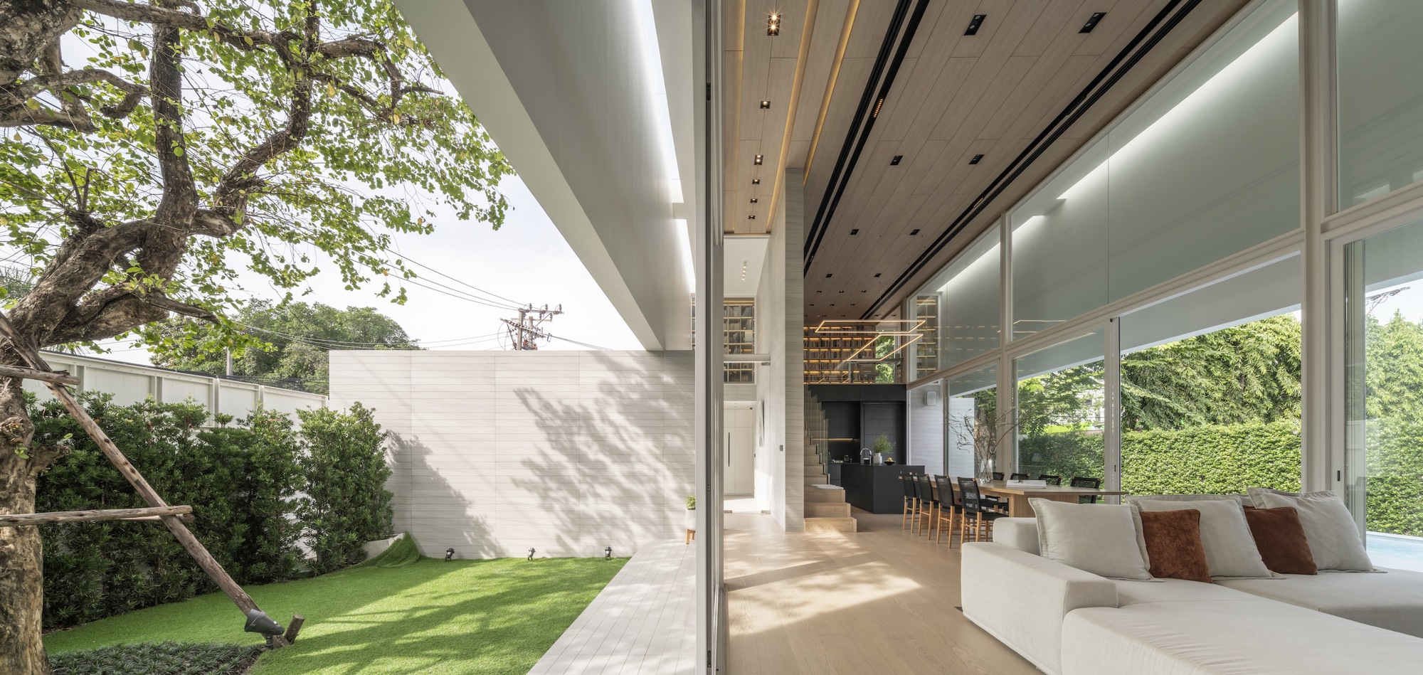 View through glass walls to the side courtyard with lawn and dappled tree shadows on white boundary wall