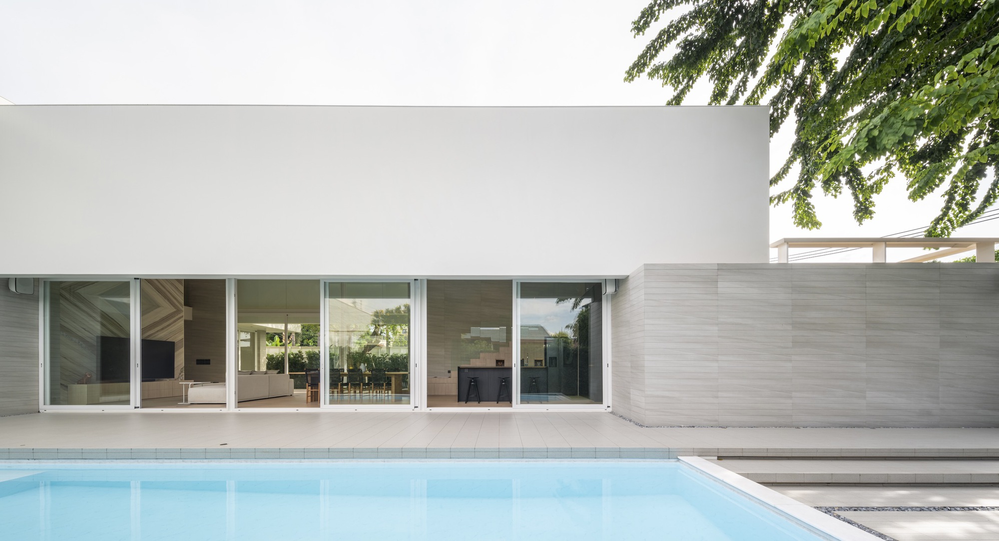 White facade with sliding glass doors opening to a pool terrace under overhanging tree branches