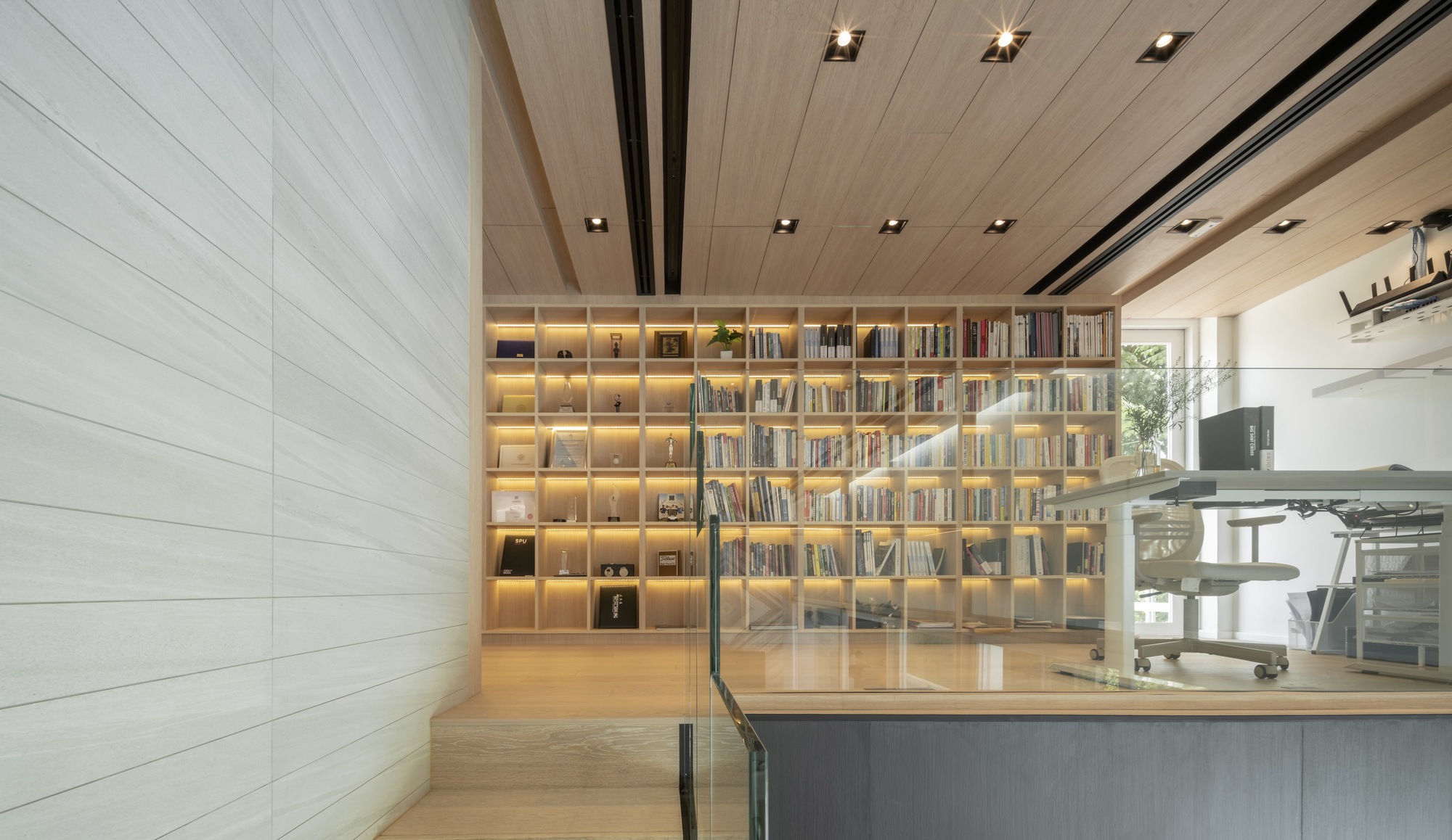 Interior view of backlit bookshelf wall with timber ceiling and glass balustrade overlooking kitchen