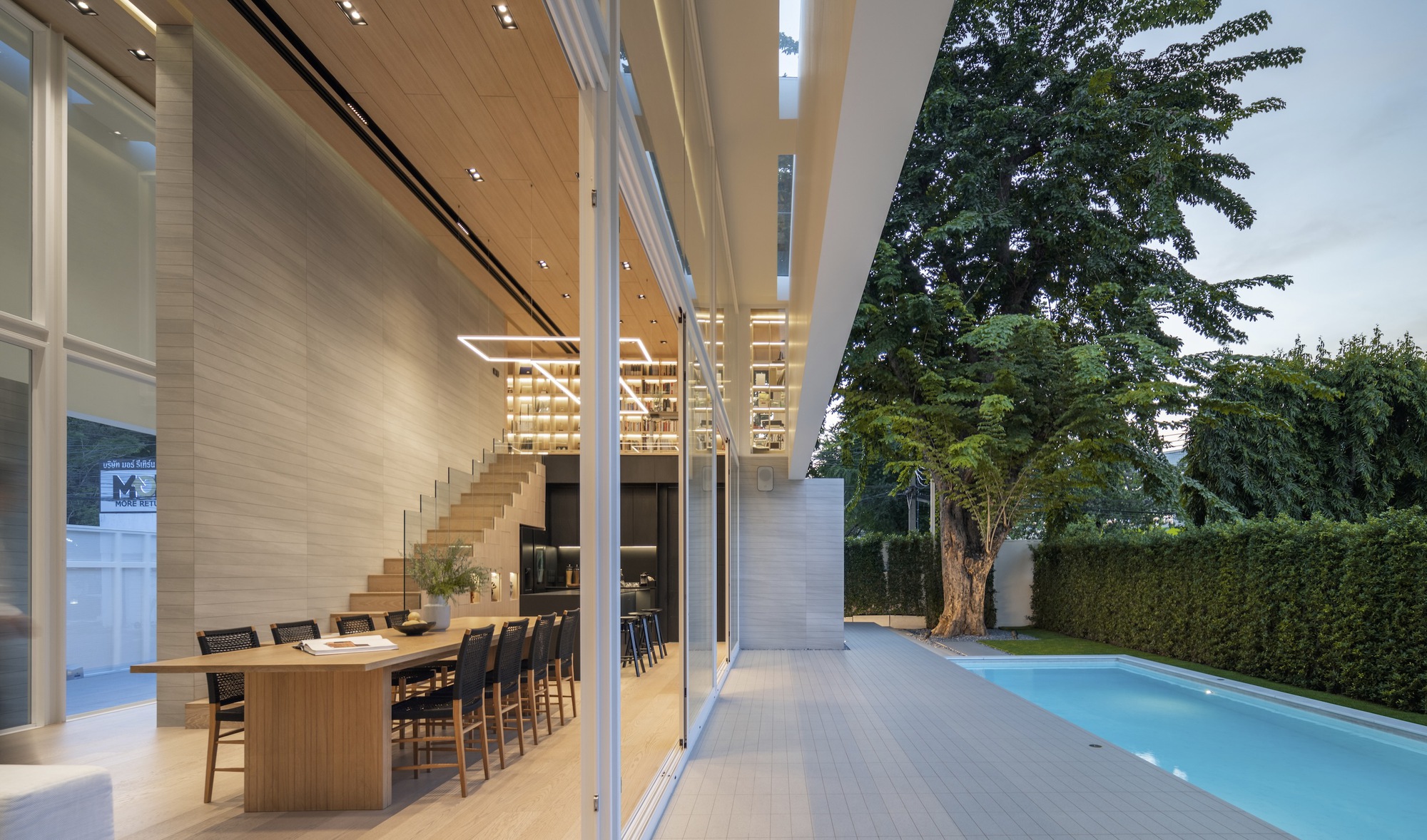 Interior view through floor-to-ceiling glass doors showing timber-lined dining space and pool deck at dusk