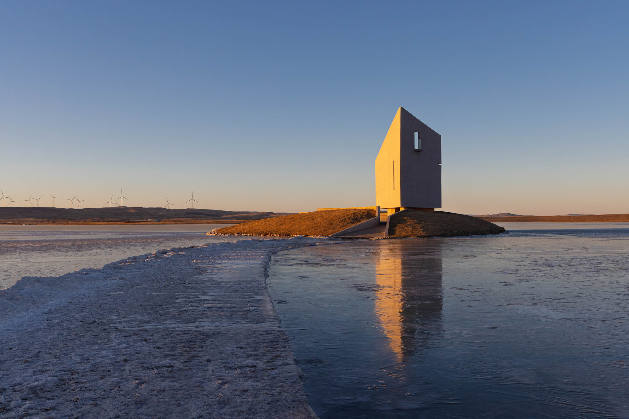 The two-tone facade reflected in the icy water with wind turbines on the horizon
