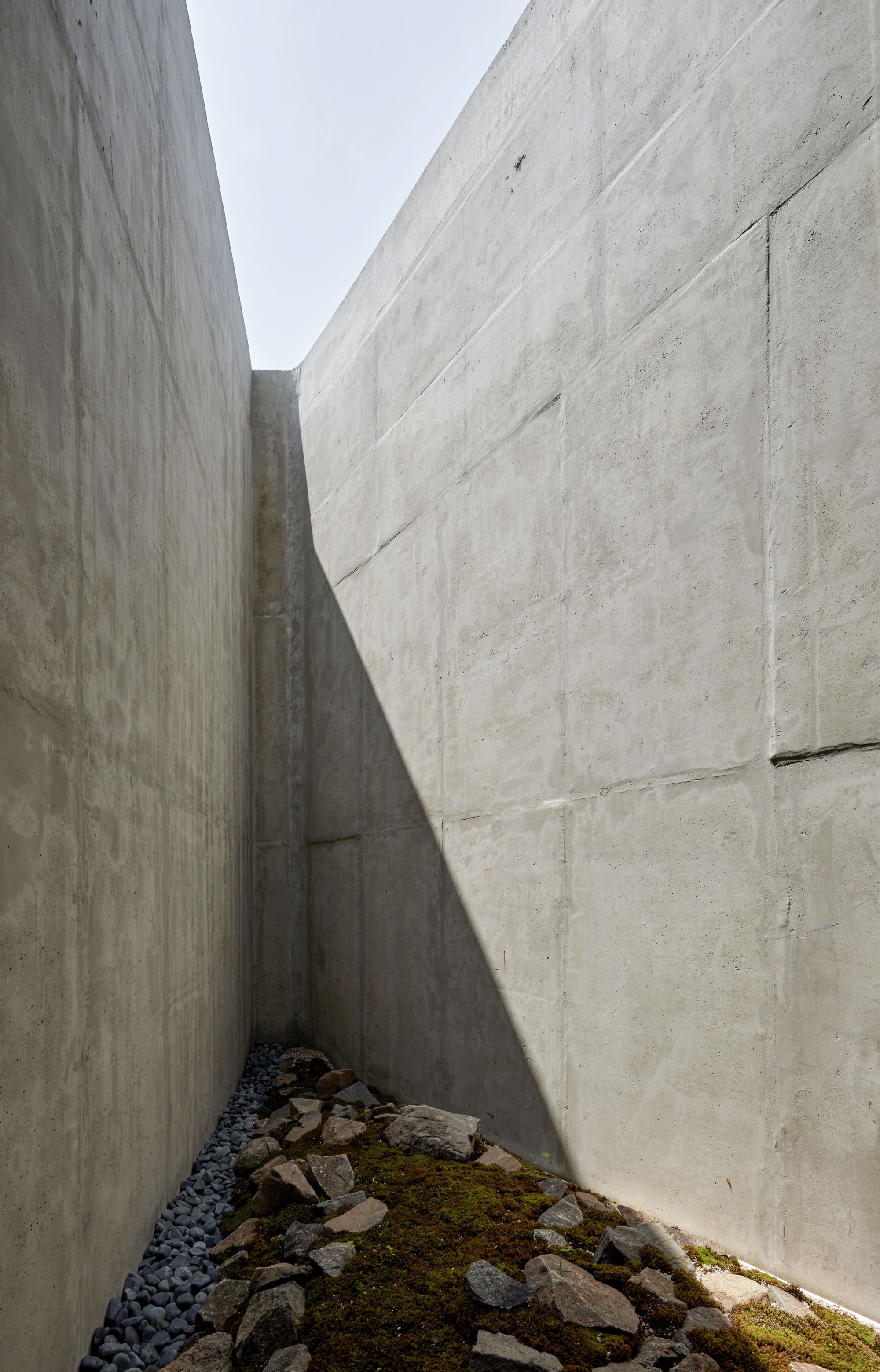 Narrow exterior courtyard between board-formed concrete walls with rocks and moss on the ground
