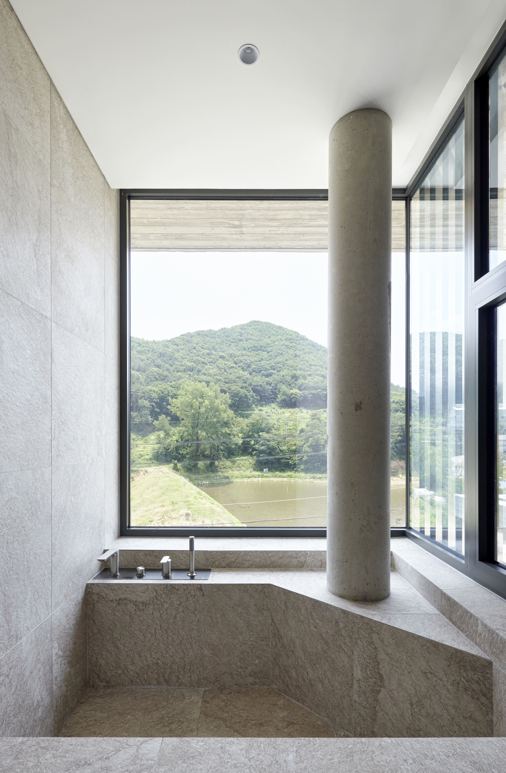 Stone bathtub with cylindrical concrete column beside a corner window framing a tree and distant mountain