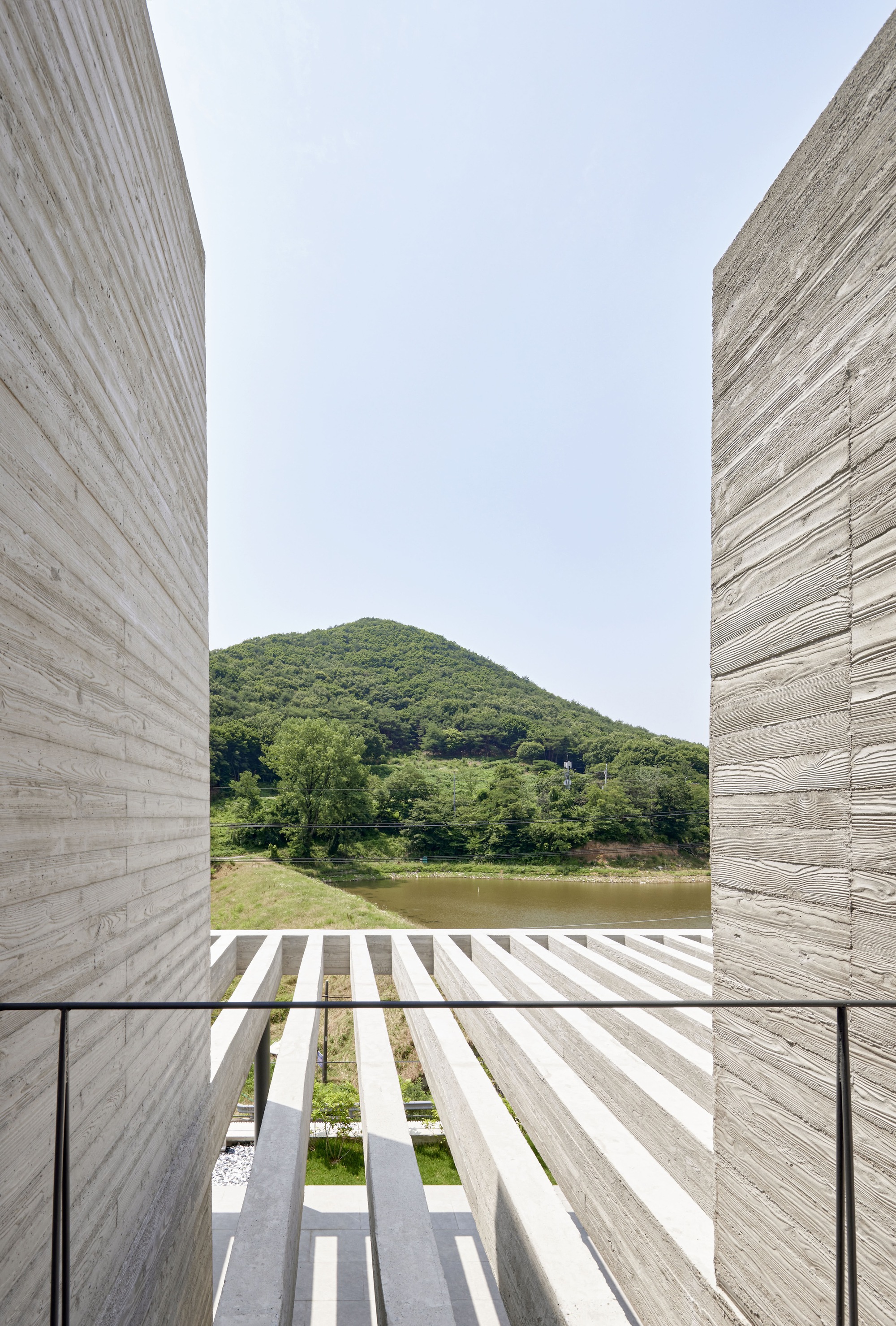 View through board-formed concrete walls toward a slatted timber deck, pond, and green hills beyond
