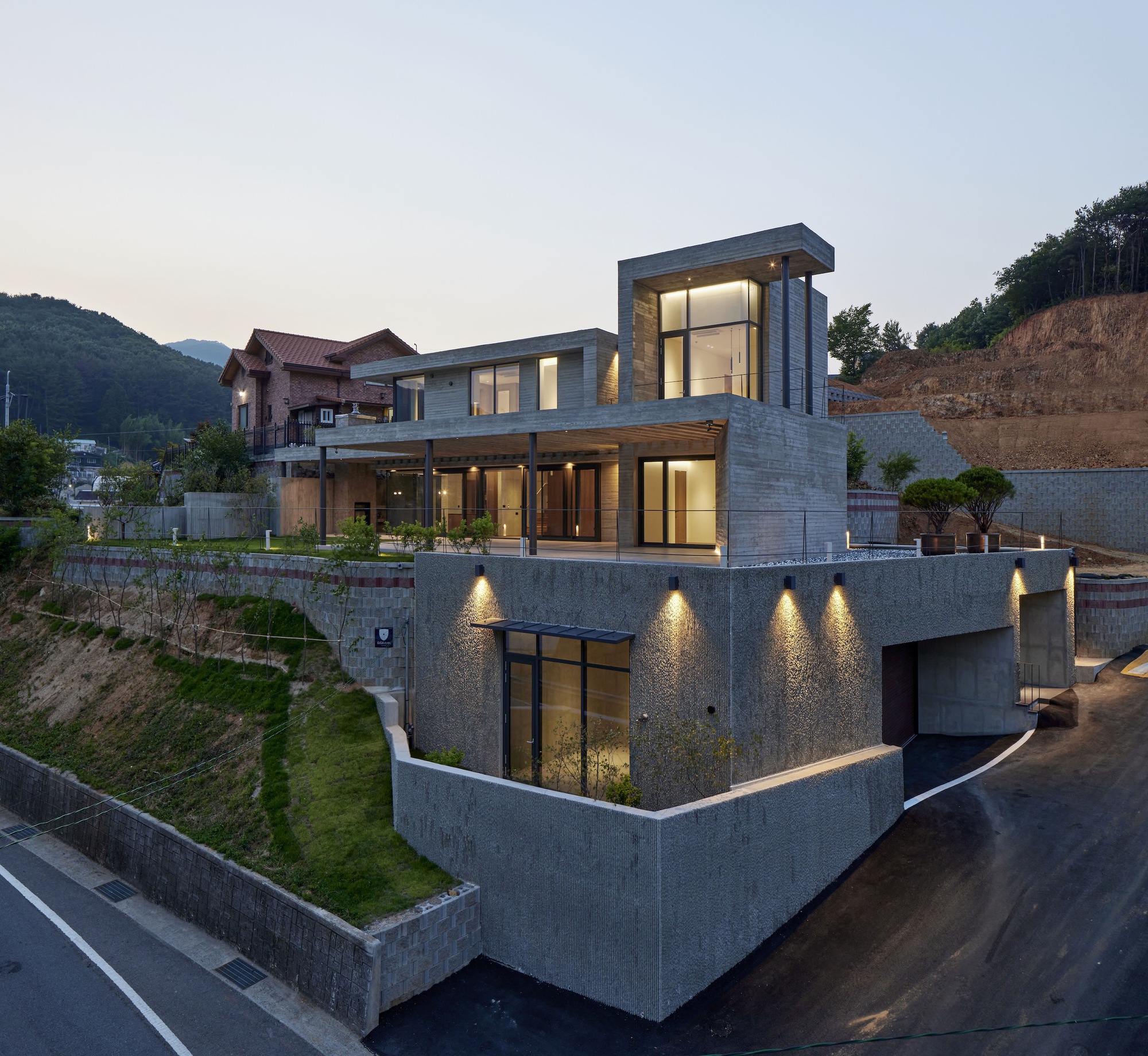Three-level concrete residence on sloped site with glazed openings illuminated at dusk