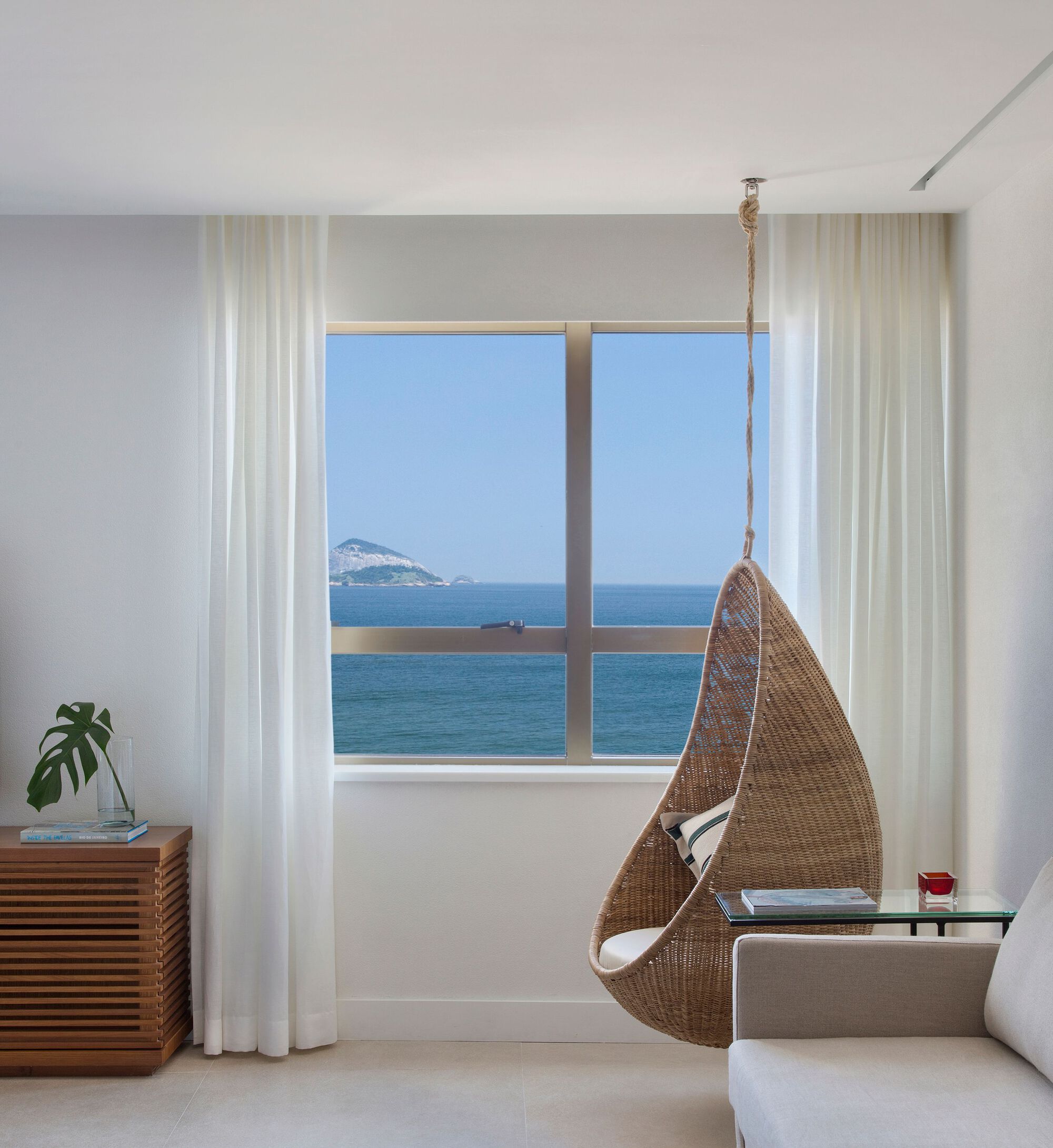Window seat nook with suspended wicker chair and ocean view through sheer white curtains
