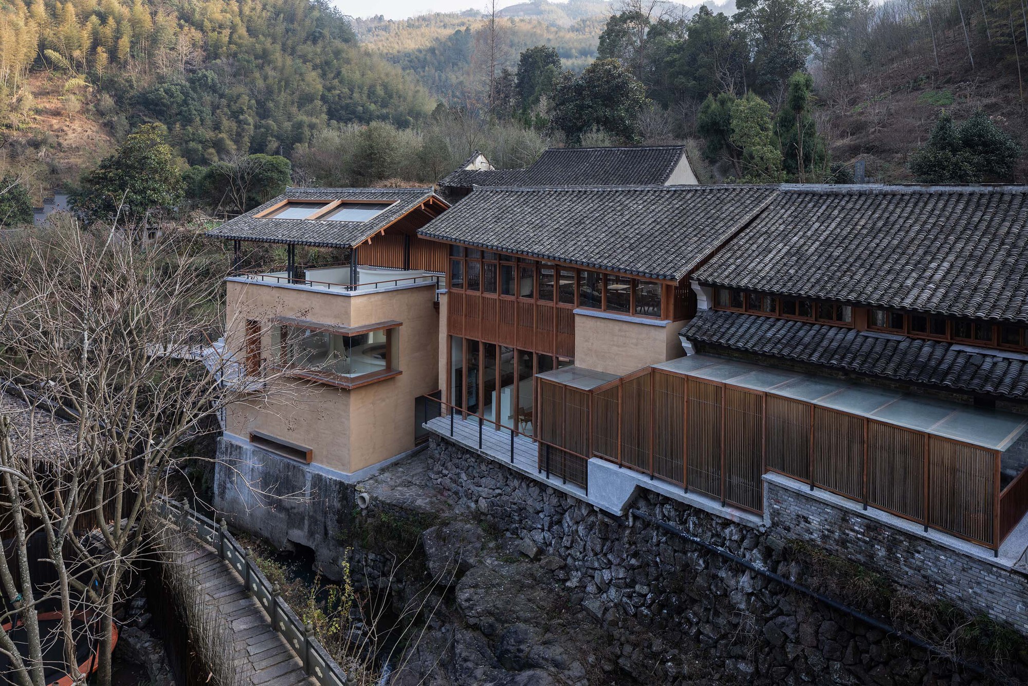 Elevated view showing timber and stucco volumes atop a stone retaining wall among bare trees
