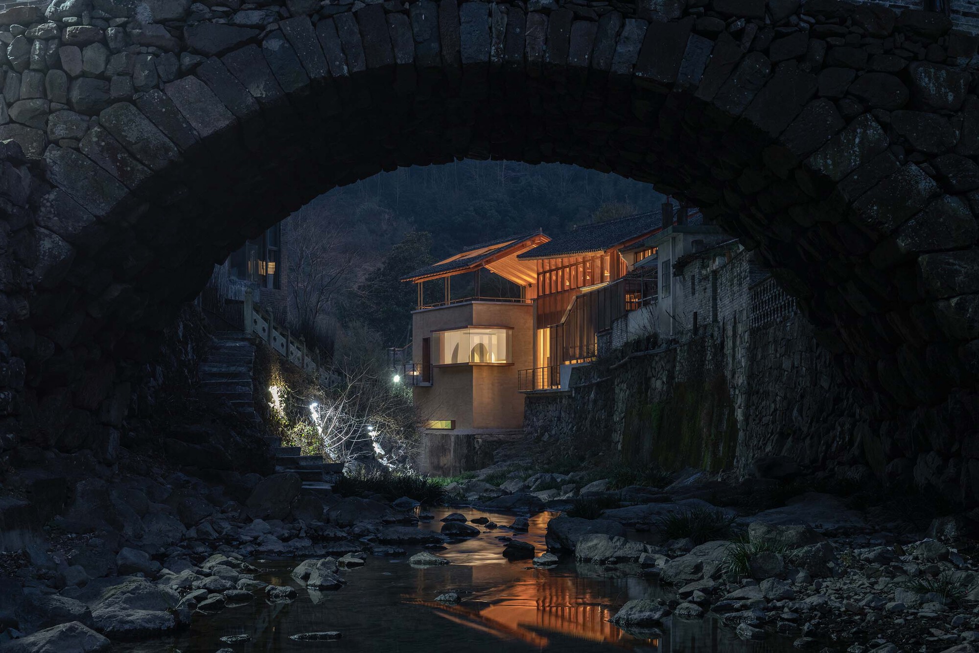 View through a stone arch bridge to the timber-clad facade illuminated at dusk above a rocky stream