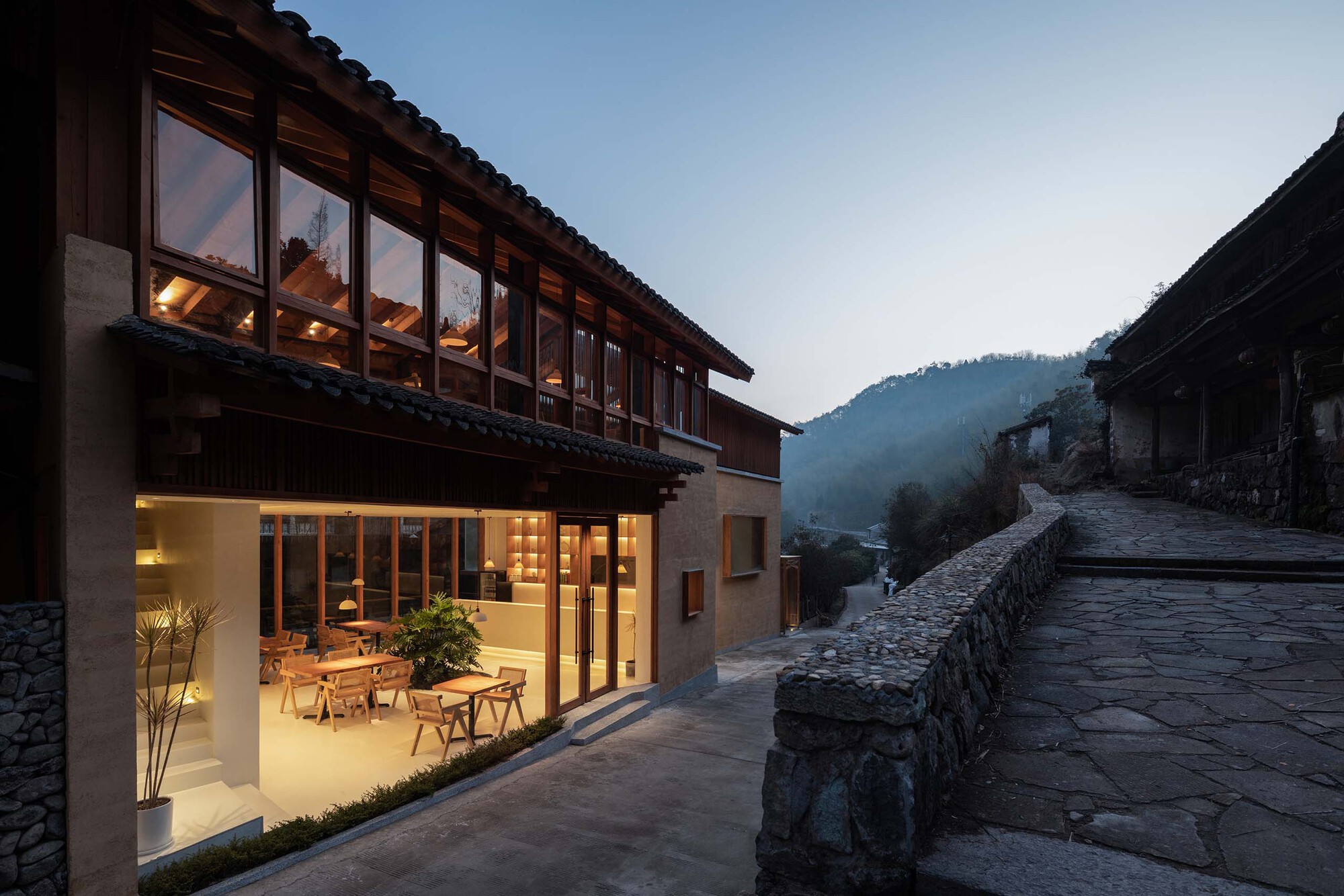 Two-story facade with recessed terrace and full-height windows opening onto flagstone courtyard in morning mist
