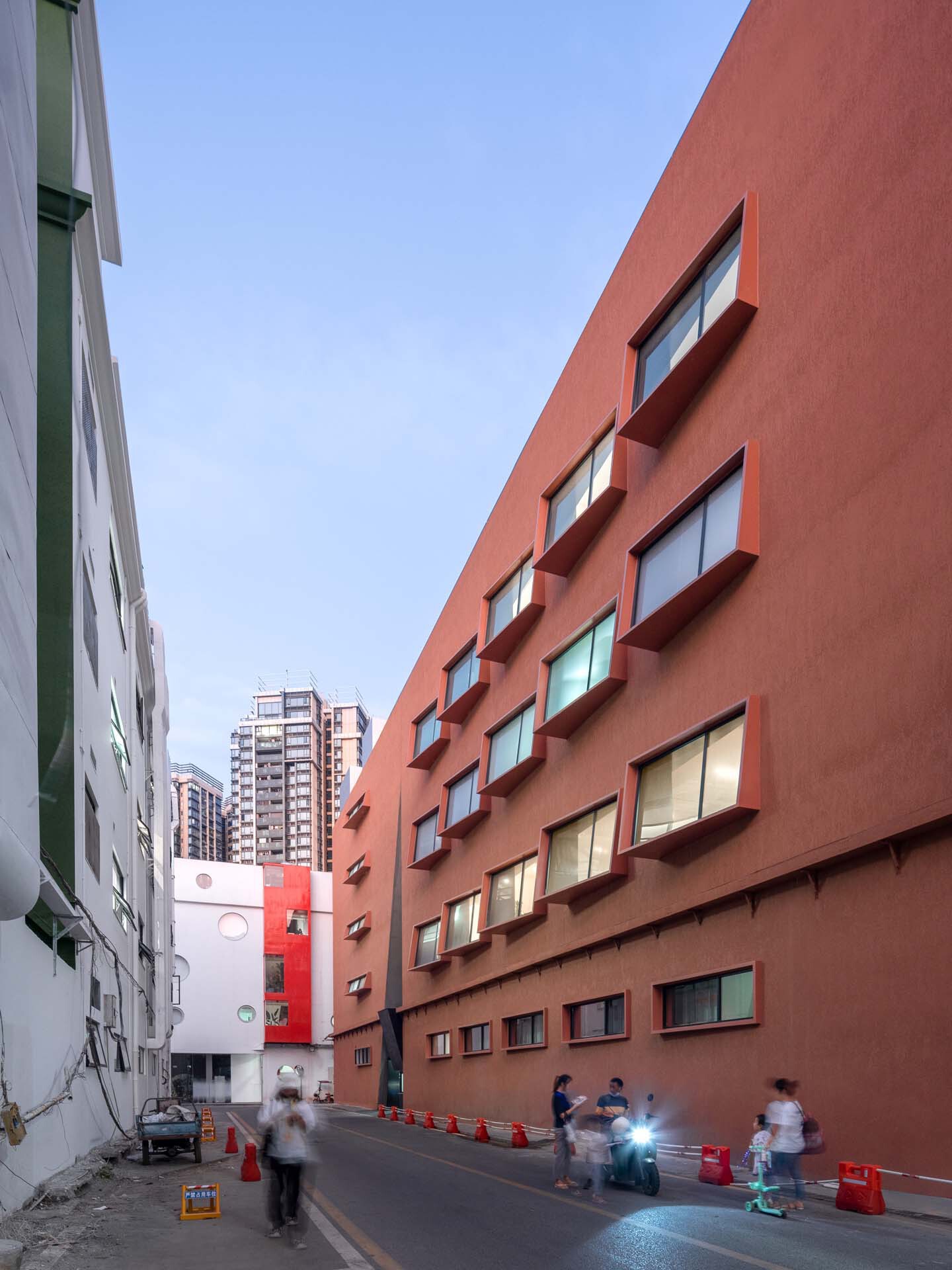 The terracotta facade with recessed windows at dusk with pedestrians and scooters passing through the alley