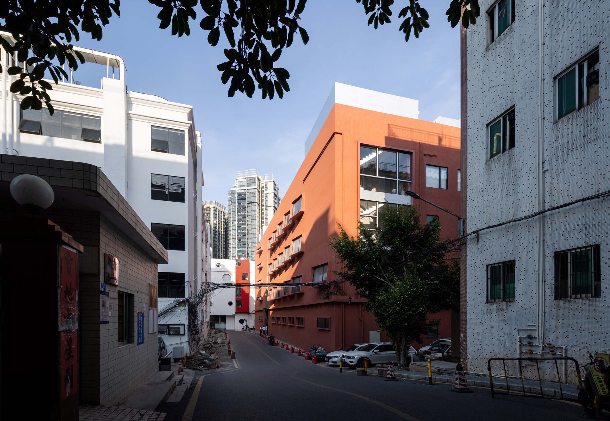 Street view of the terracotta volume nestled between white and grey buildings under overhanging tree branches