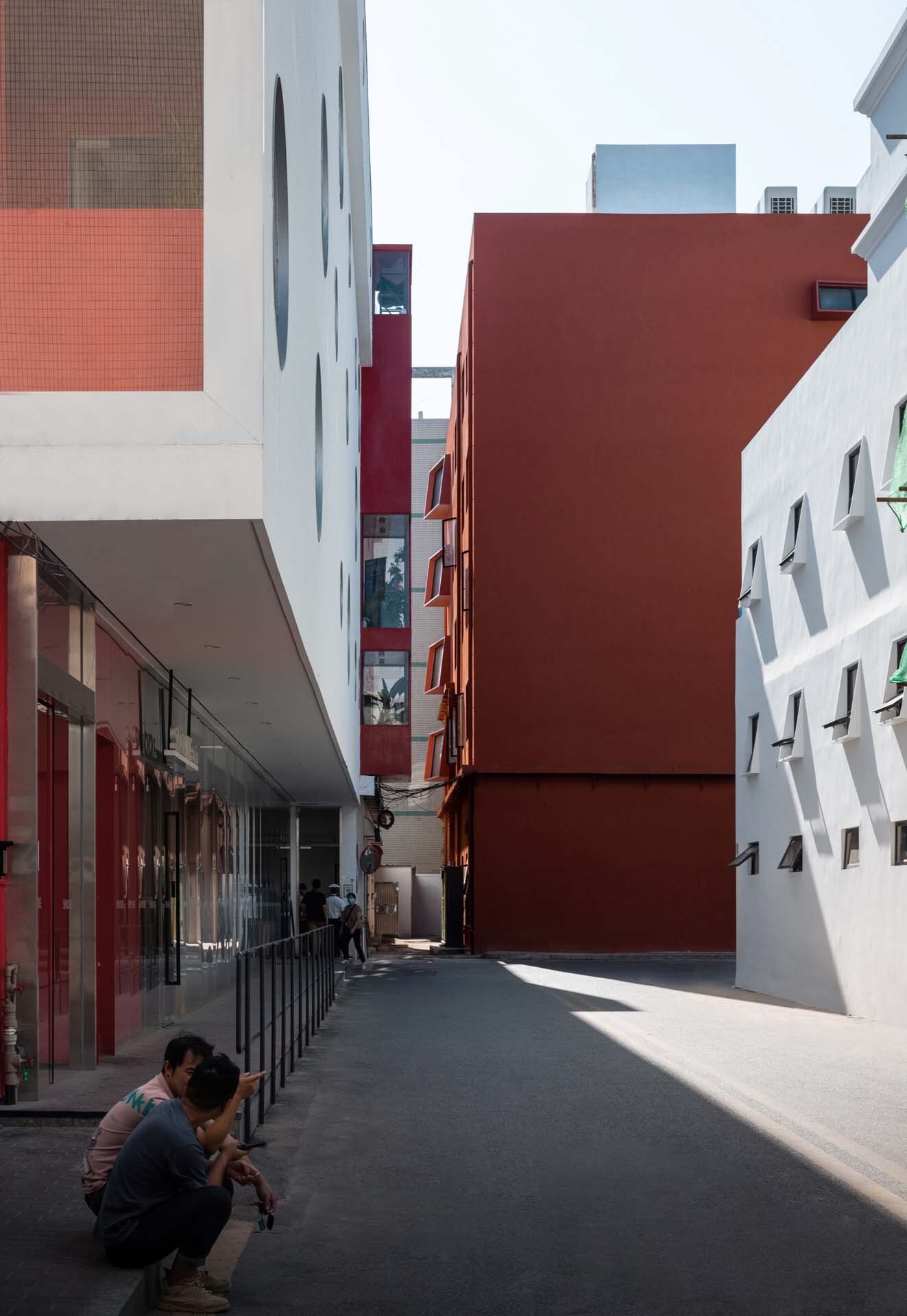 Narrow pedestrian passage between the terracotta volume and white perforated facade with a person sitting in shadow