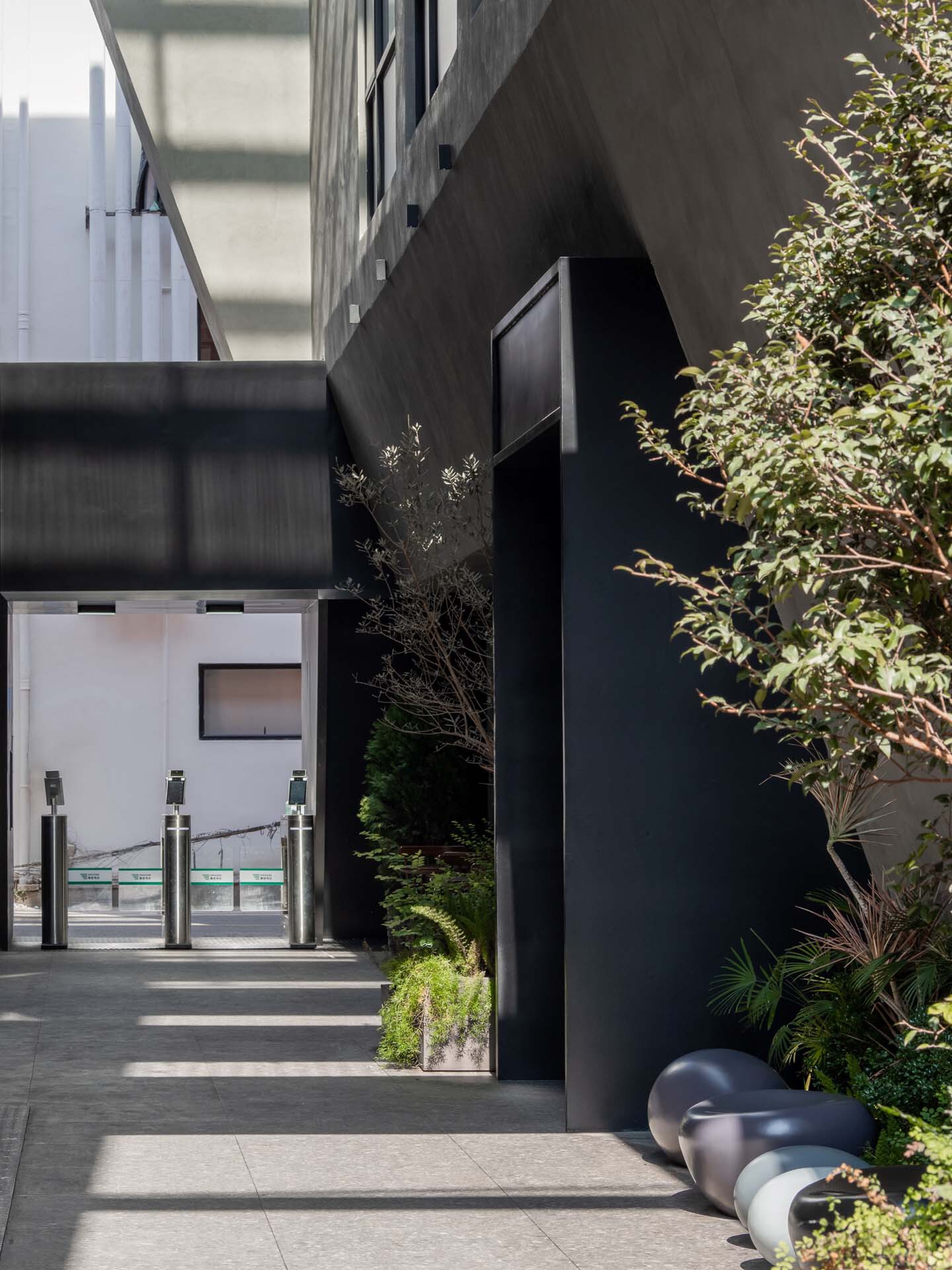 Courtyard entrance with black bollards, planted beds and a glazed security gate in midday sun