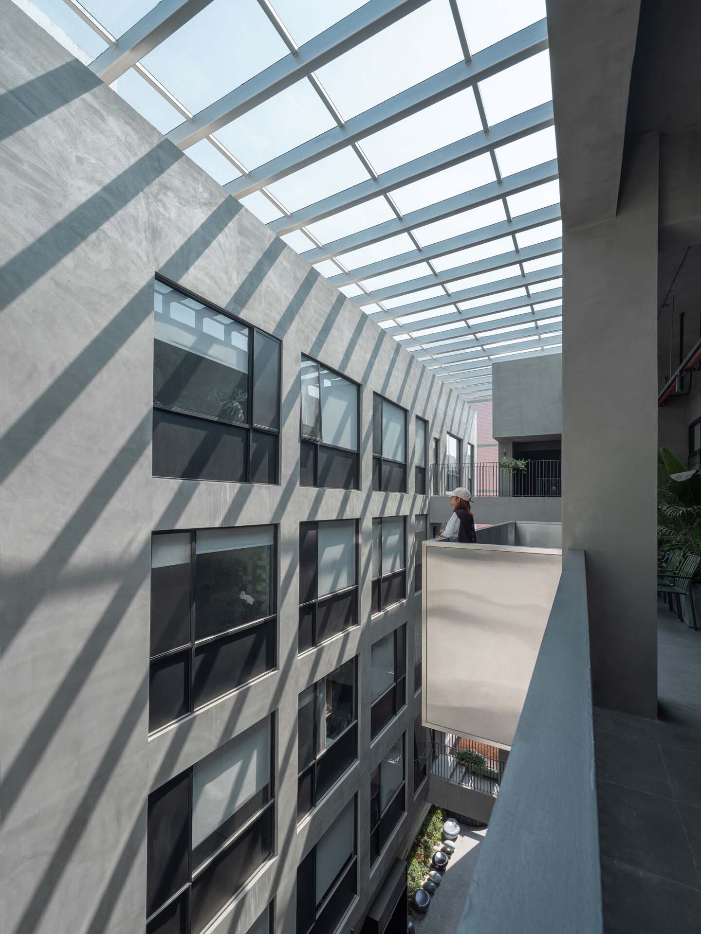 Interior courtyard balcony with diagonal shadows from overhead glazed skylight and gridded windows along the wall