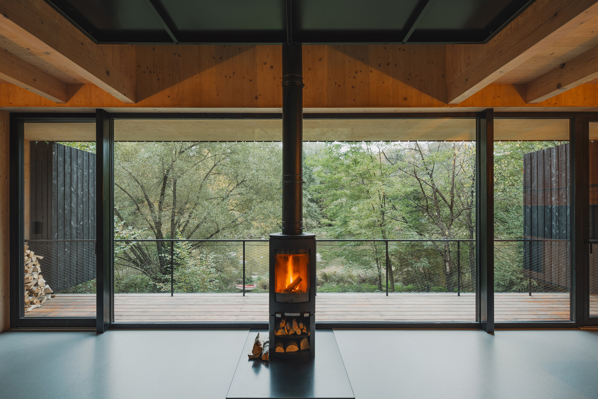 Freestanding wood stove centered before full-height glazing overlooking forest in afternoon light