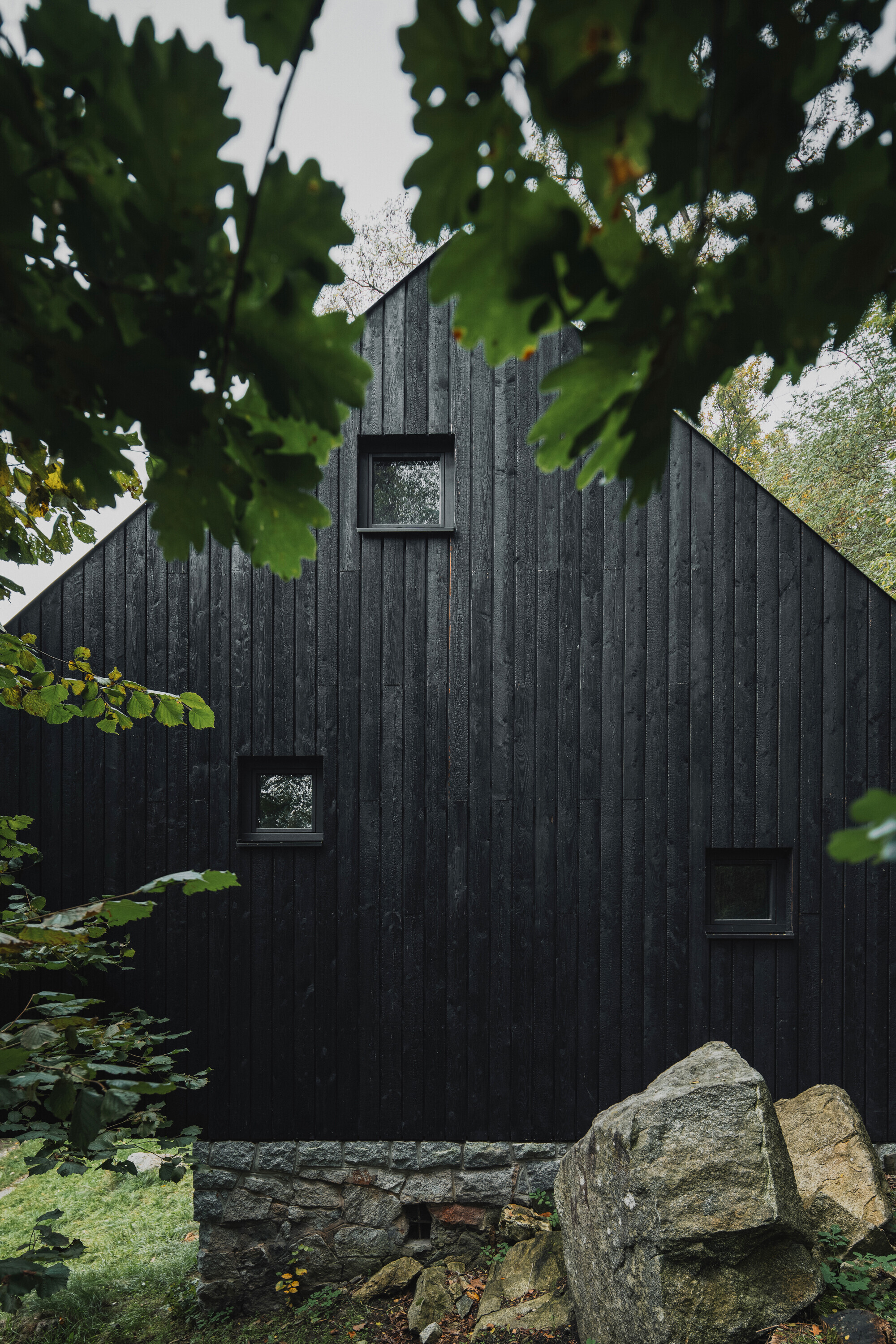 Charred timber facade with small square windows on a stone foundation beneath leafy branches