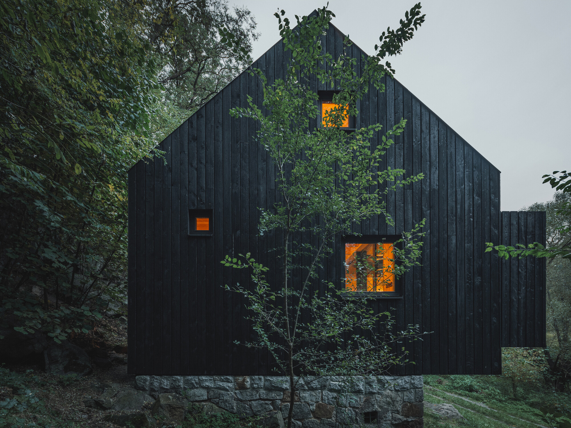 Black vertical timber cladding on a gabled volume with illuminated window openings at dusk