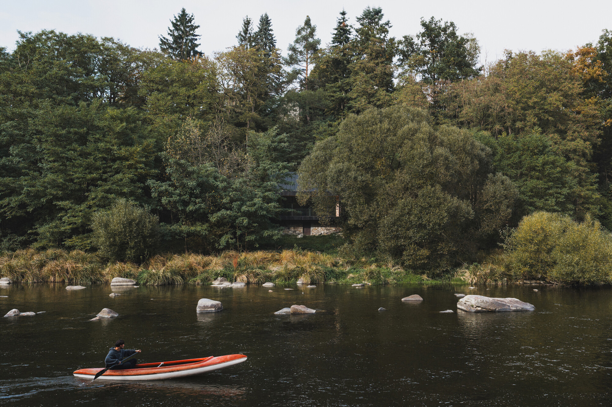 Distant view from the river showing the structure nestled among dense trees with a kayak