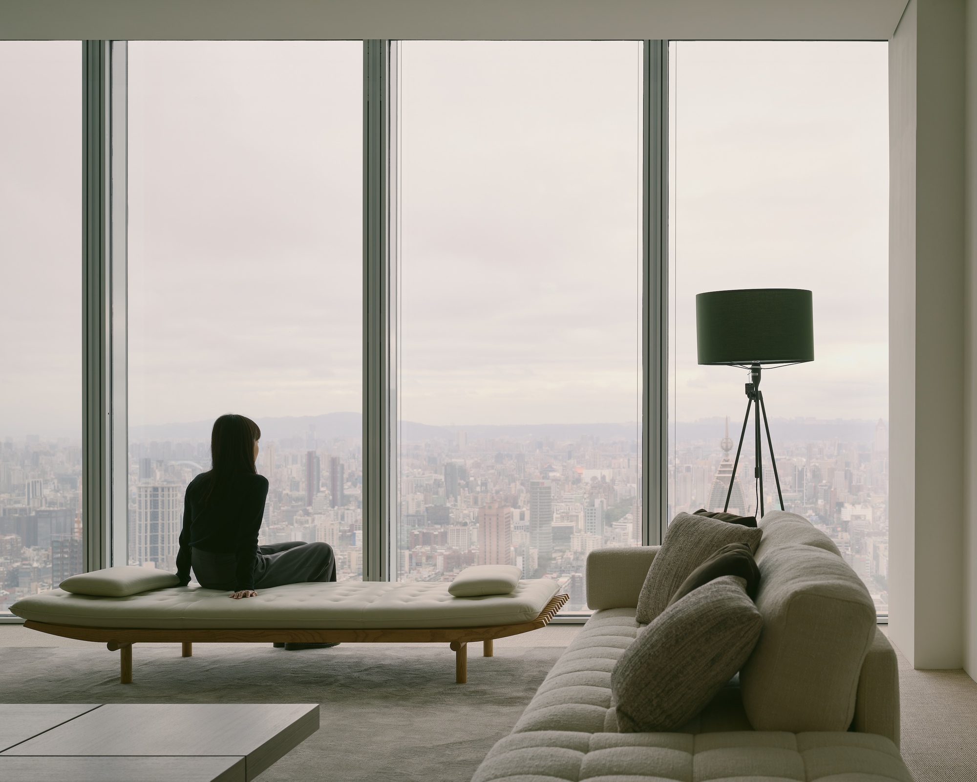 Person sitting on daybed by full-height windows overlooking the cityscape in soft afternoon light