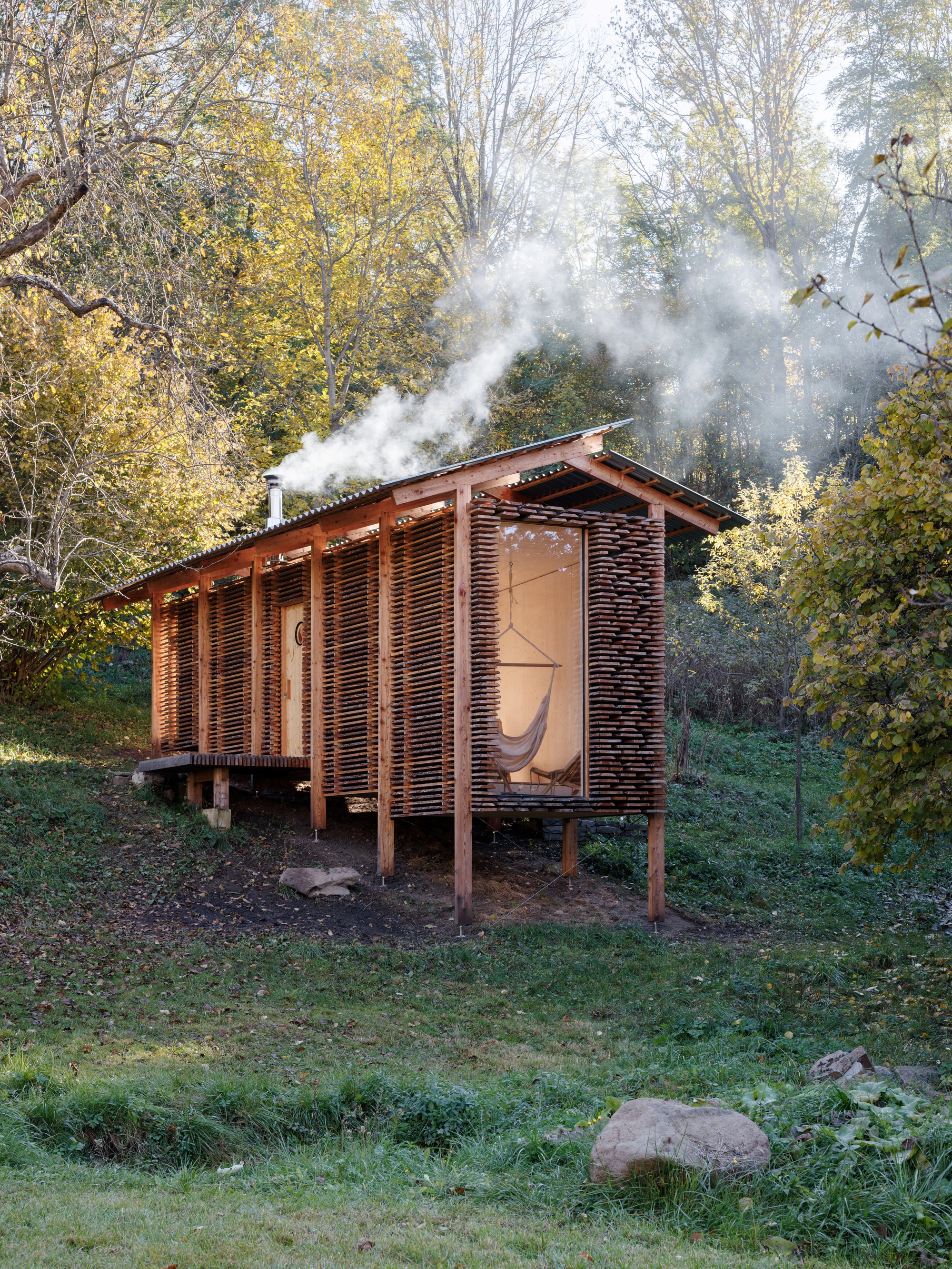 Elevated timber pavilion with slatted walls and pitched metal roof with chimney smoke rising at dusk