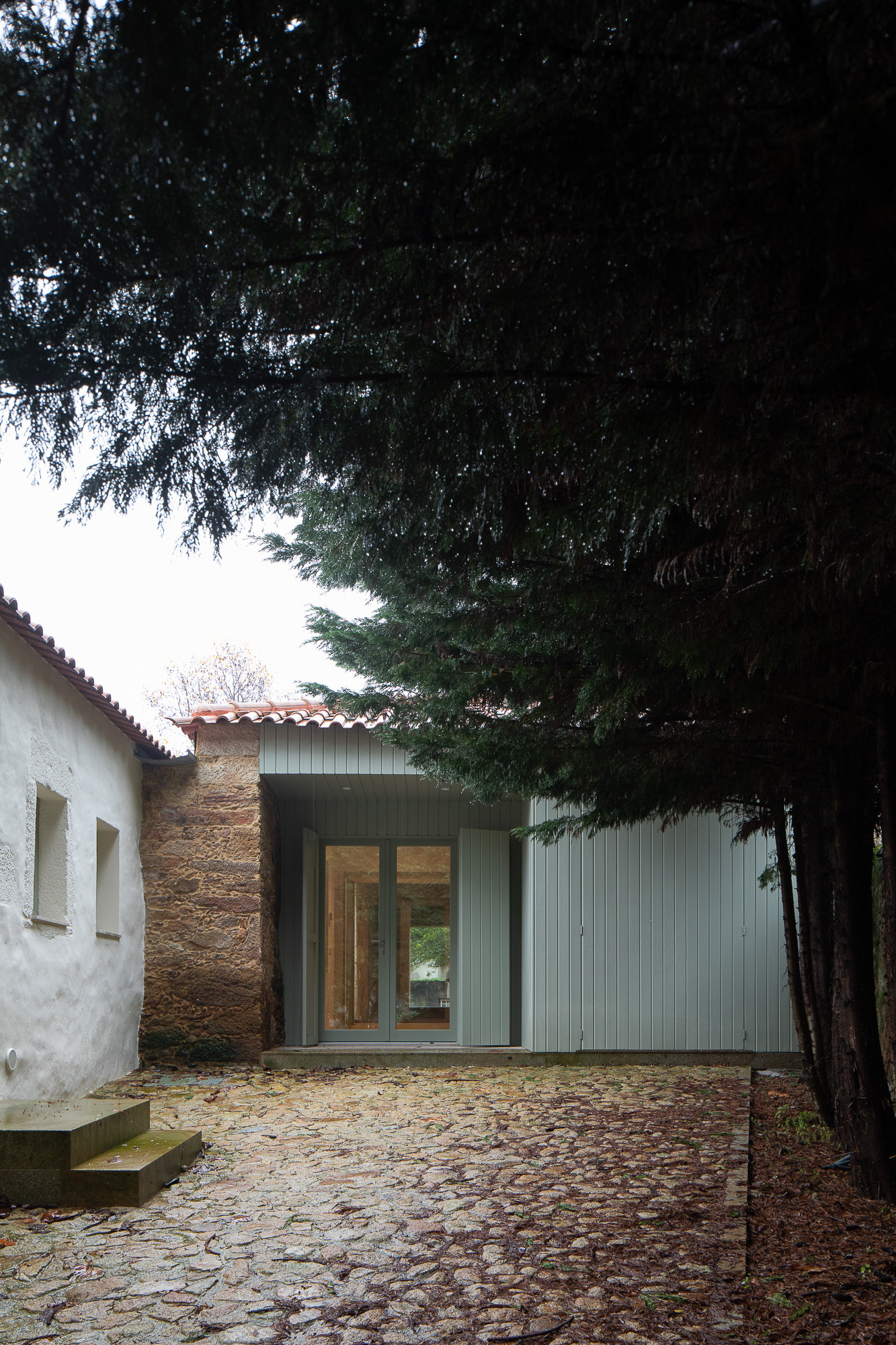 Corrugated metal pavilion inserted between whitewashed wall and stone structure under evergreen canopy
