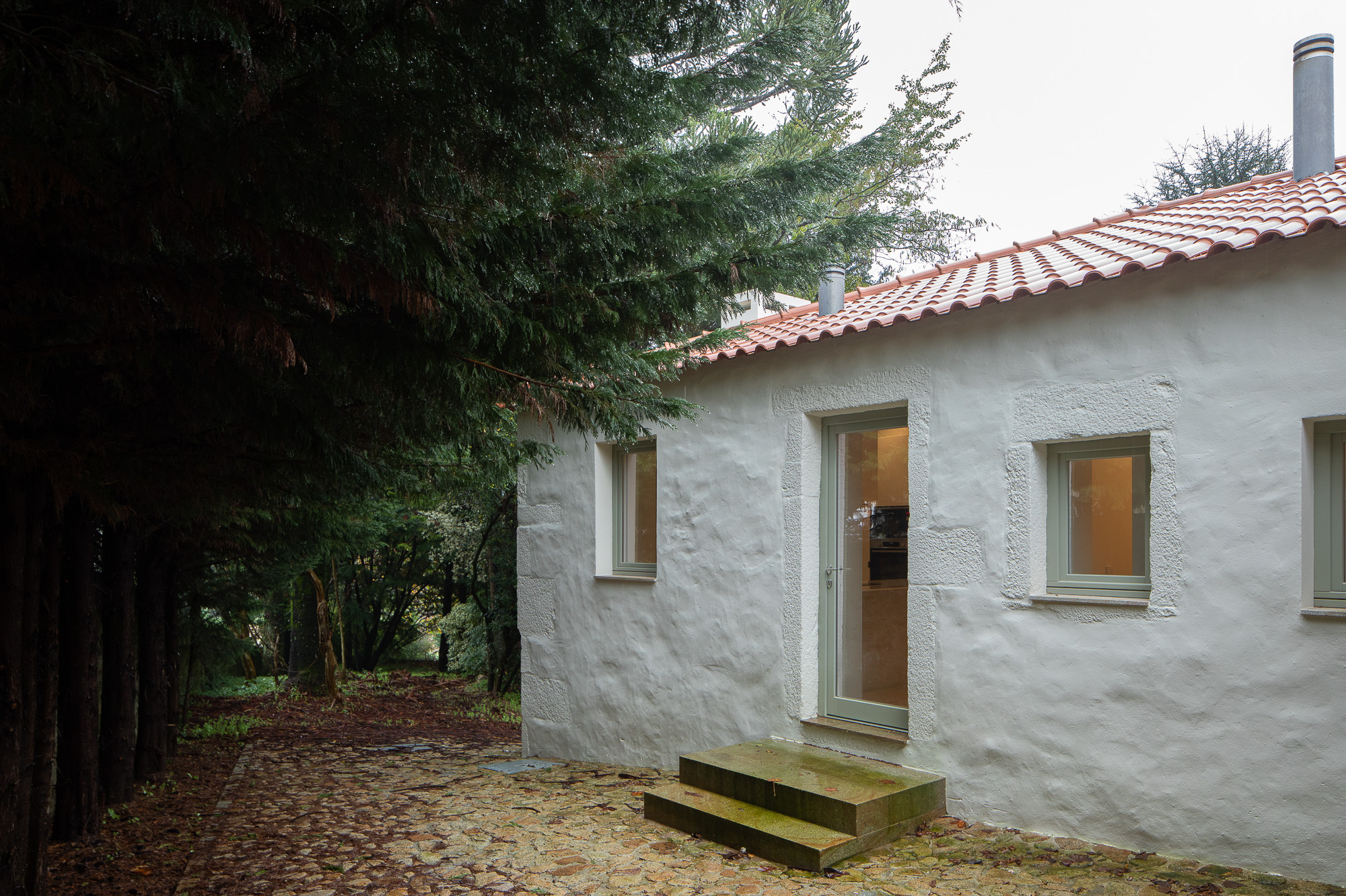 White rendered facade with timber-framed windows and terracotta tile roof beside evergreen trees