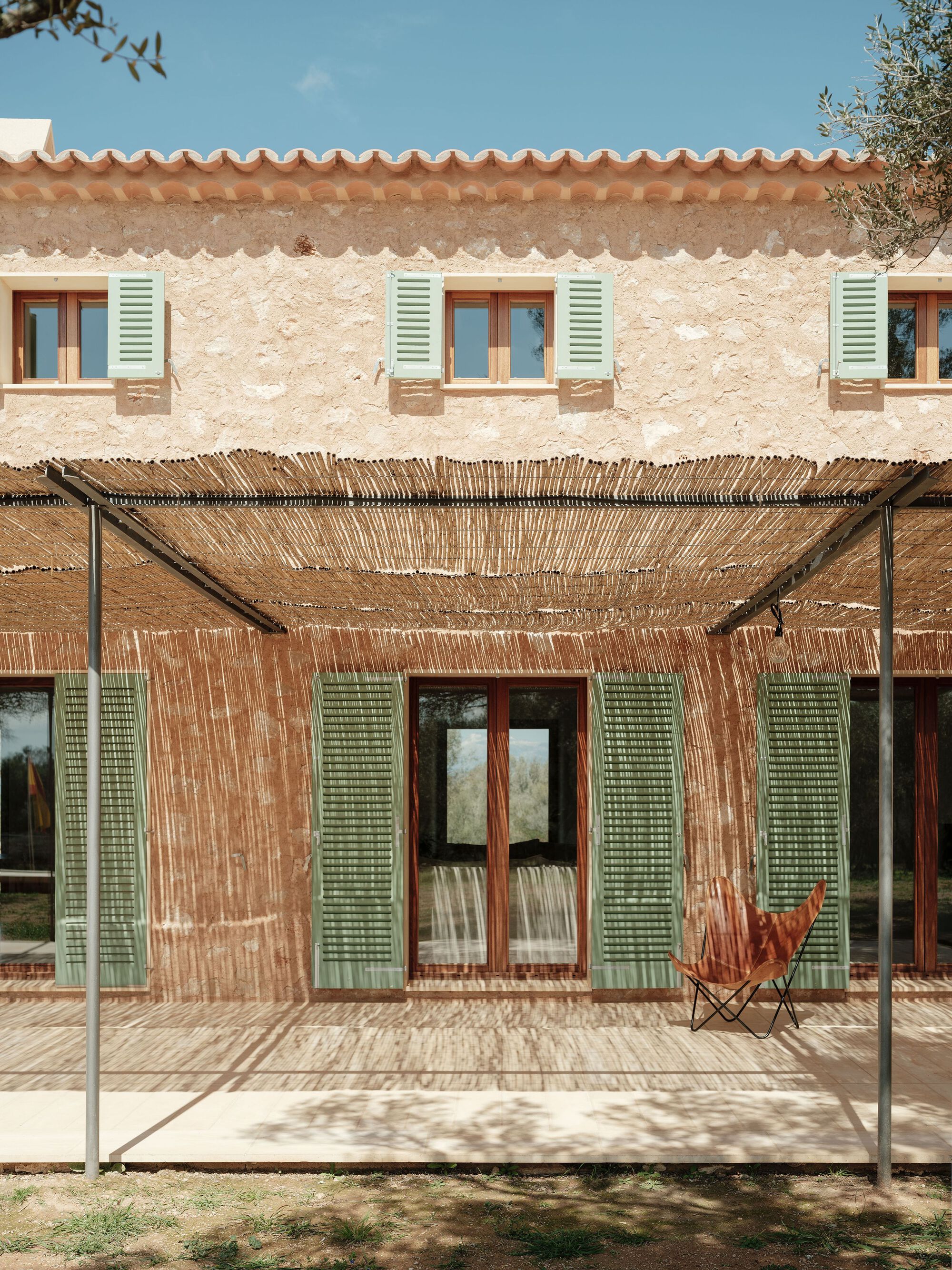 Stone facade with terra cotta tile roof and reed pergola shading glass doors and green shutters