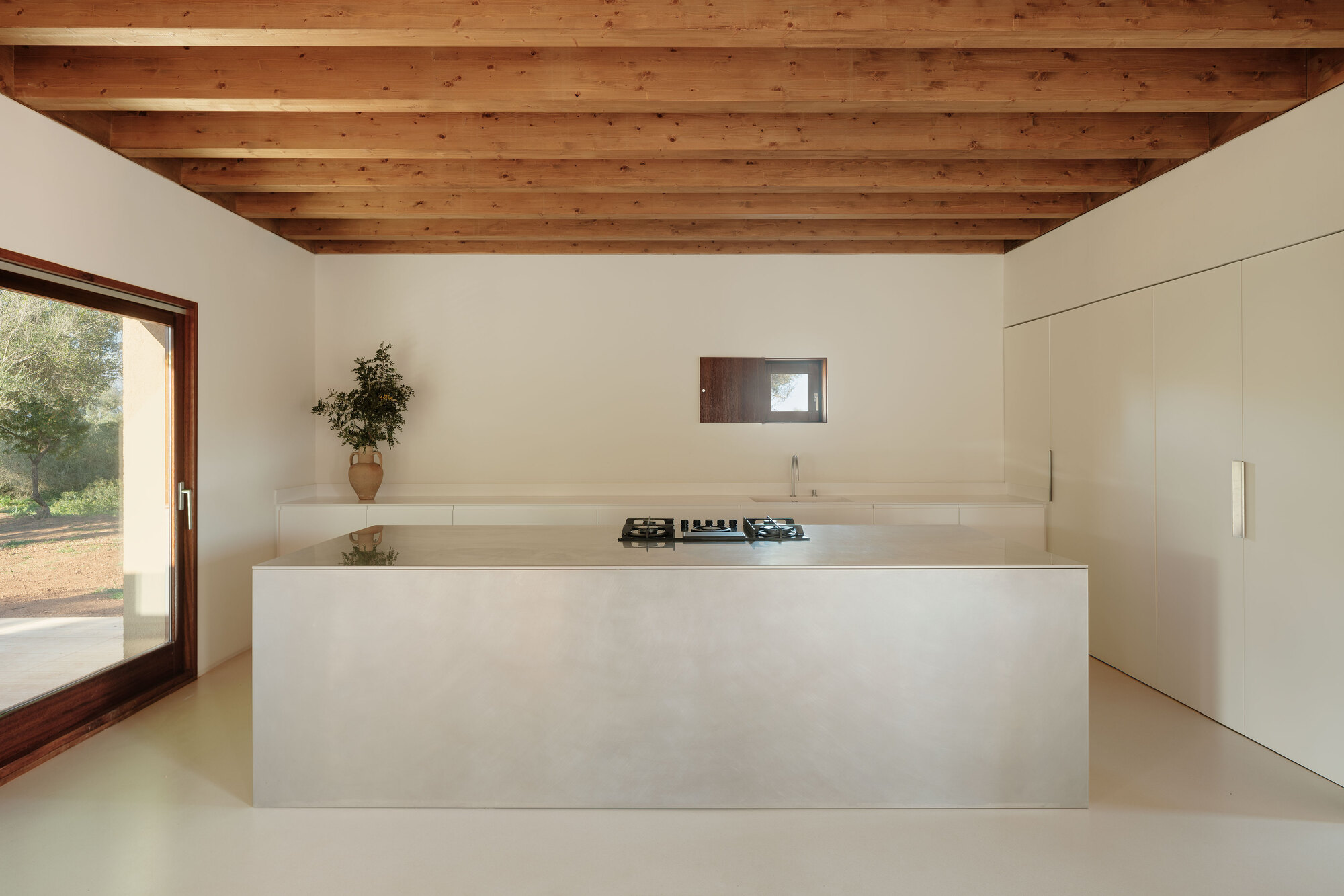 Kitchen island with integrated cooktop beneath timber ceiling and small horizontal window in plastered wall