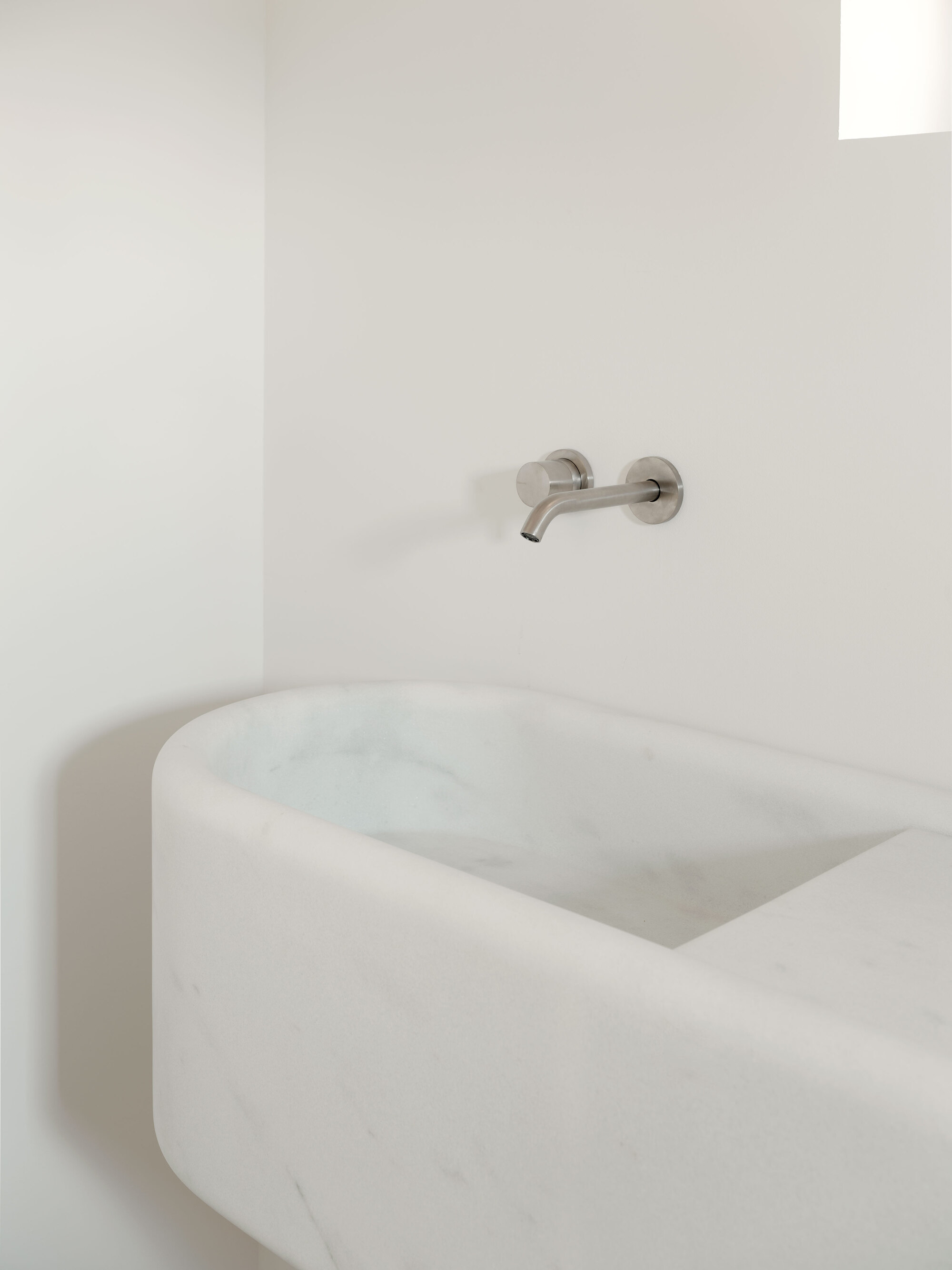 Carved marble basin with rounded edges below a wall-mounted stainless steel faucet