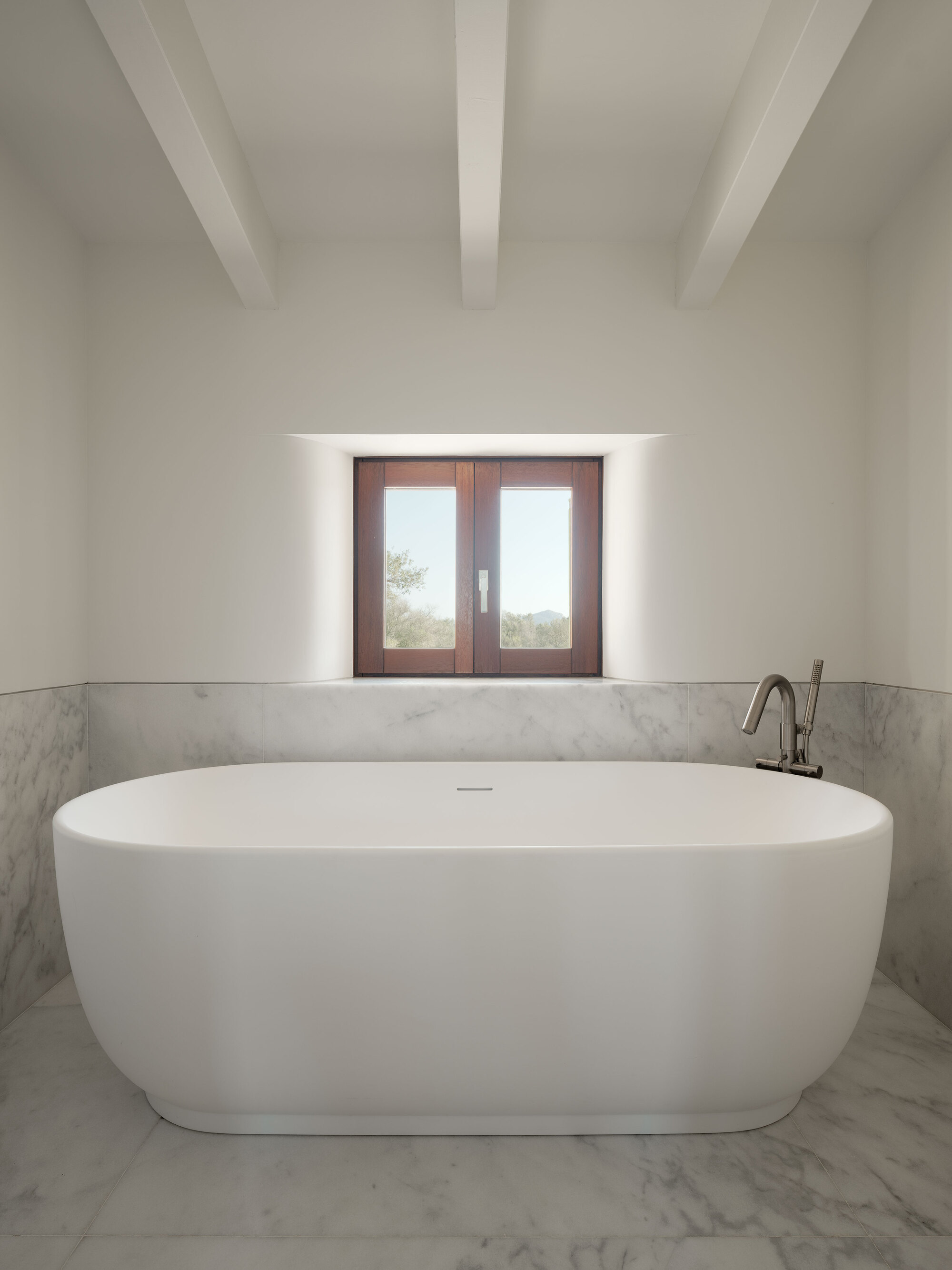Freestanding oval bathtub beneath a deep-set timber-framed window with exposed ceiling beams