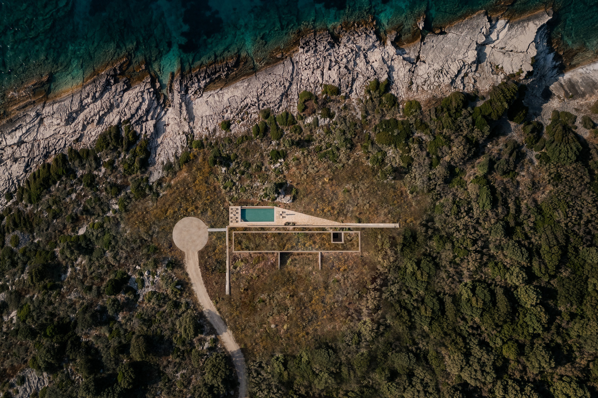 Aerial view of linear residence with swimming pool nestled between rocky coastal cliffs and dense Mediterranean vegetation