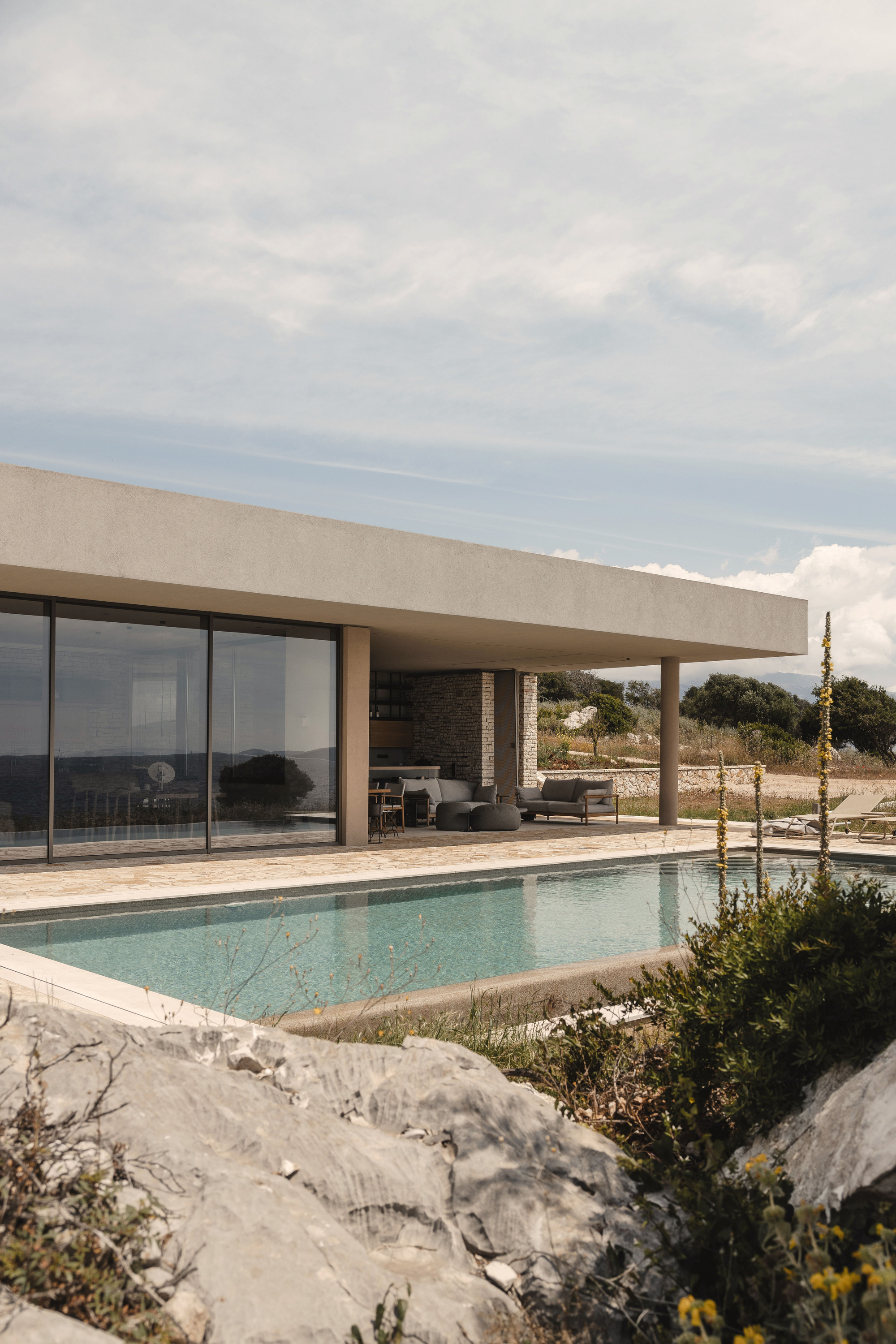 Covered terrace with stone wall and furniture overlooking rectangular pool edged by limestone outcrops and plantings
