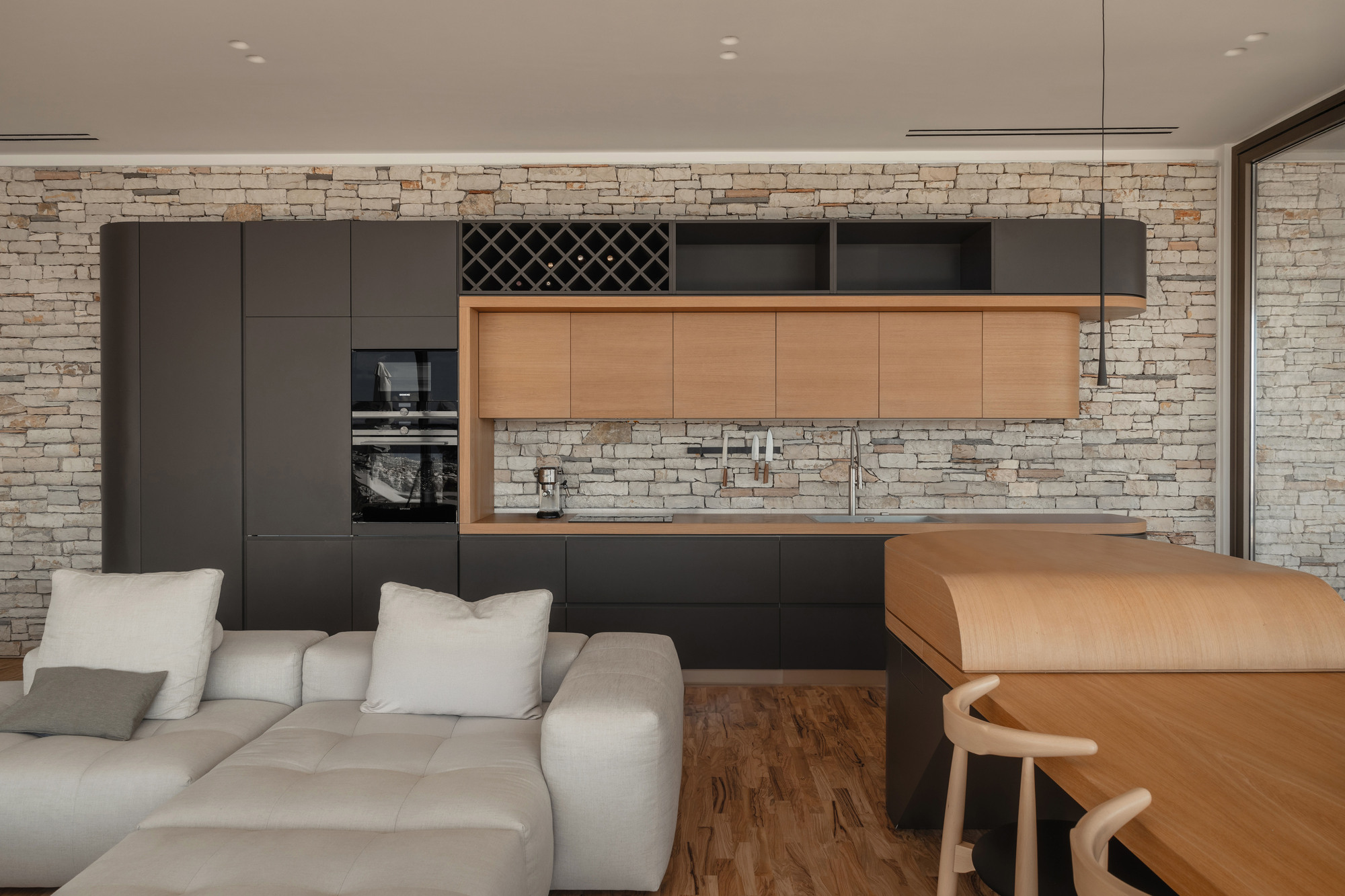 Open kitchen with black and oak cabinetry against stacked stone wall viewed from living area with sofa