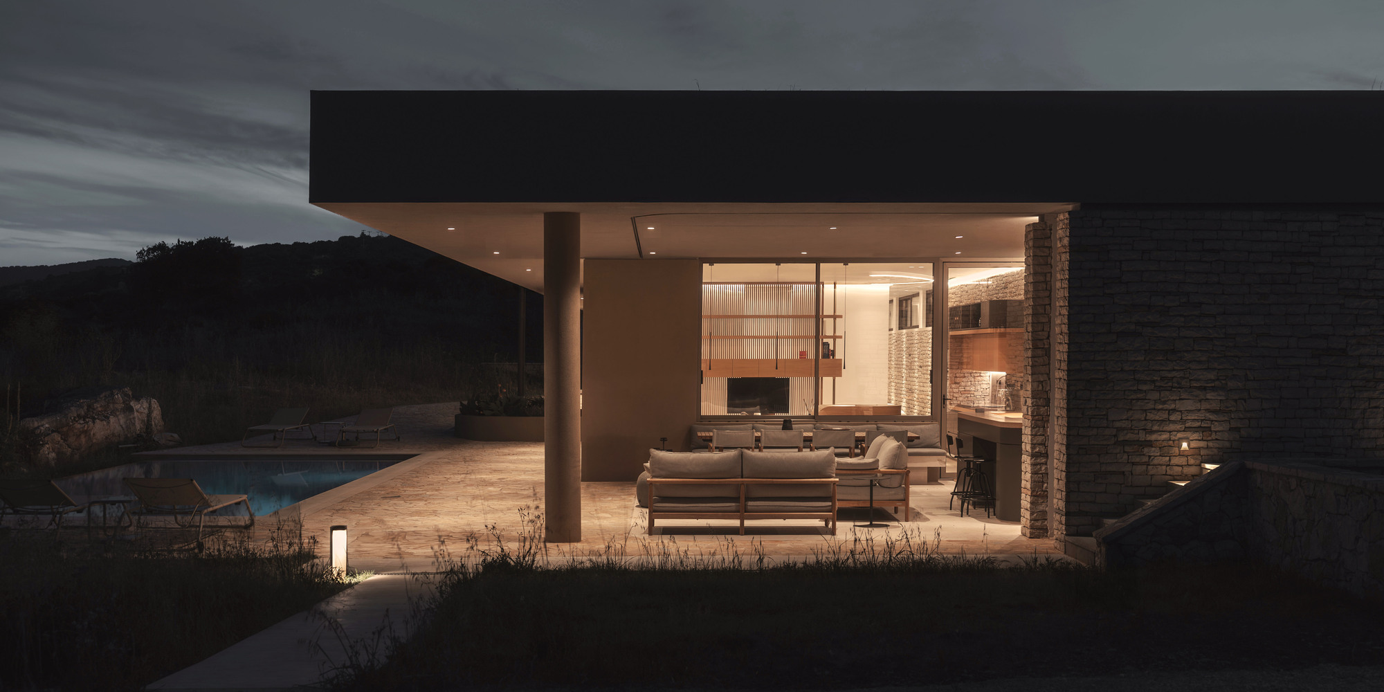 Covered outdoor living space with stacked stone fireplace illuminated at dusk overlooking the pool