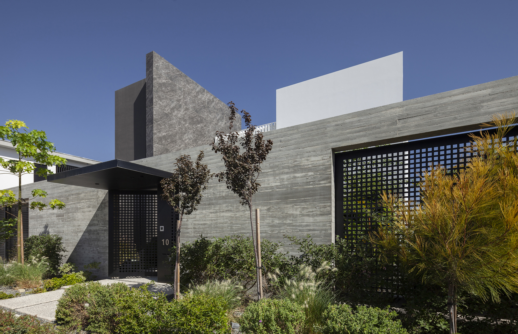 Entrance facade showing horizontal stone cladding and perforated metal screens surrounded by planted shrubs and grasses