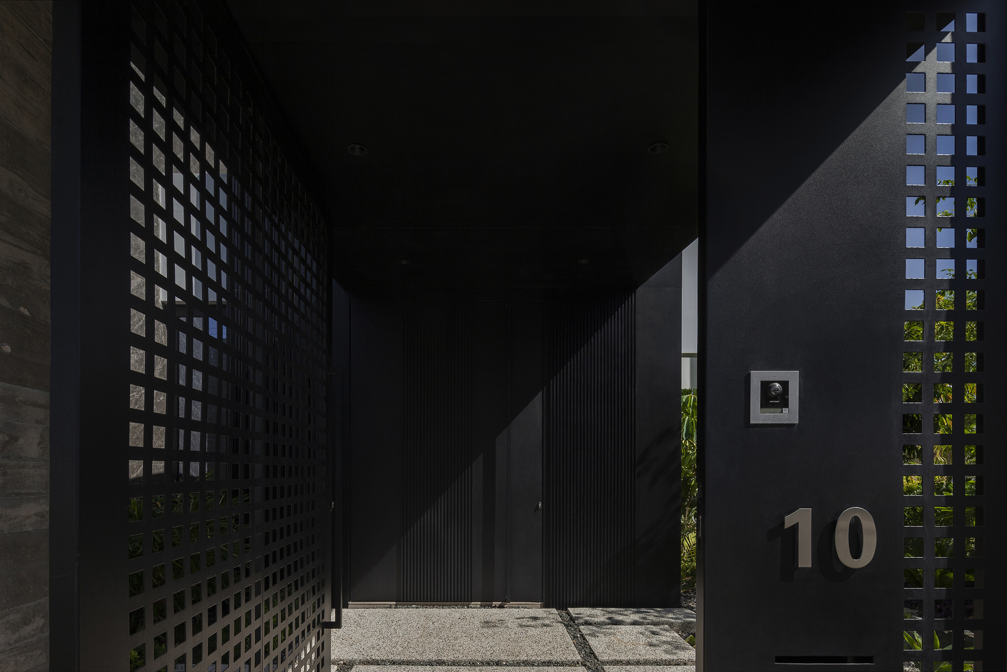 Entry courtyard with perforated black metal screens filtering sunlight onto gravel paving